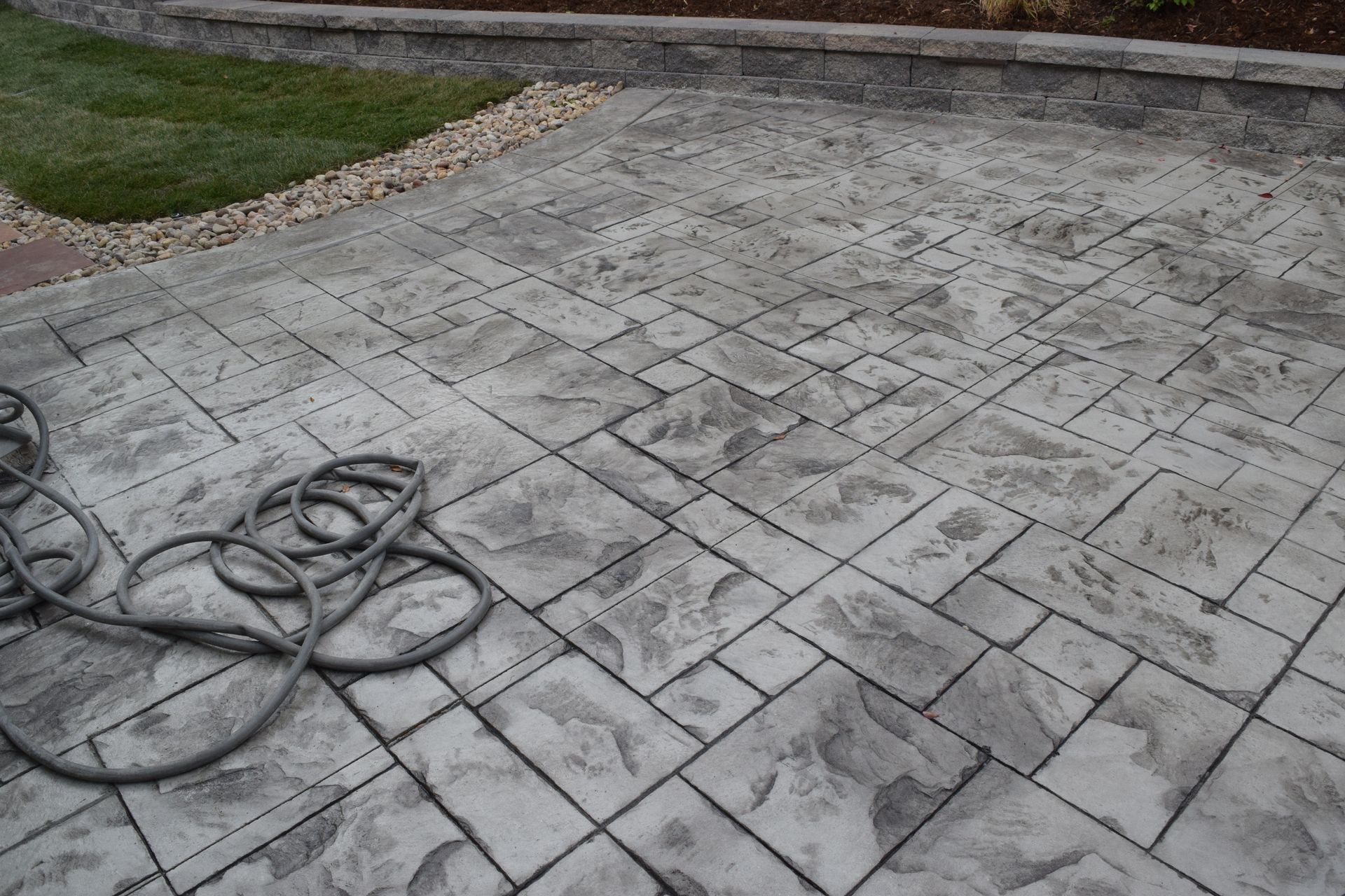 Gray stamped concrete patio with coiled hose, adjacent to a grassy area and retaining wall.