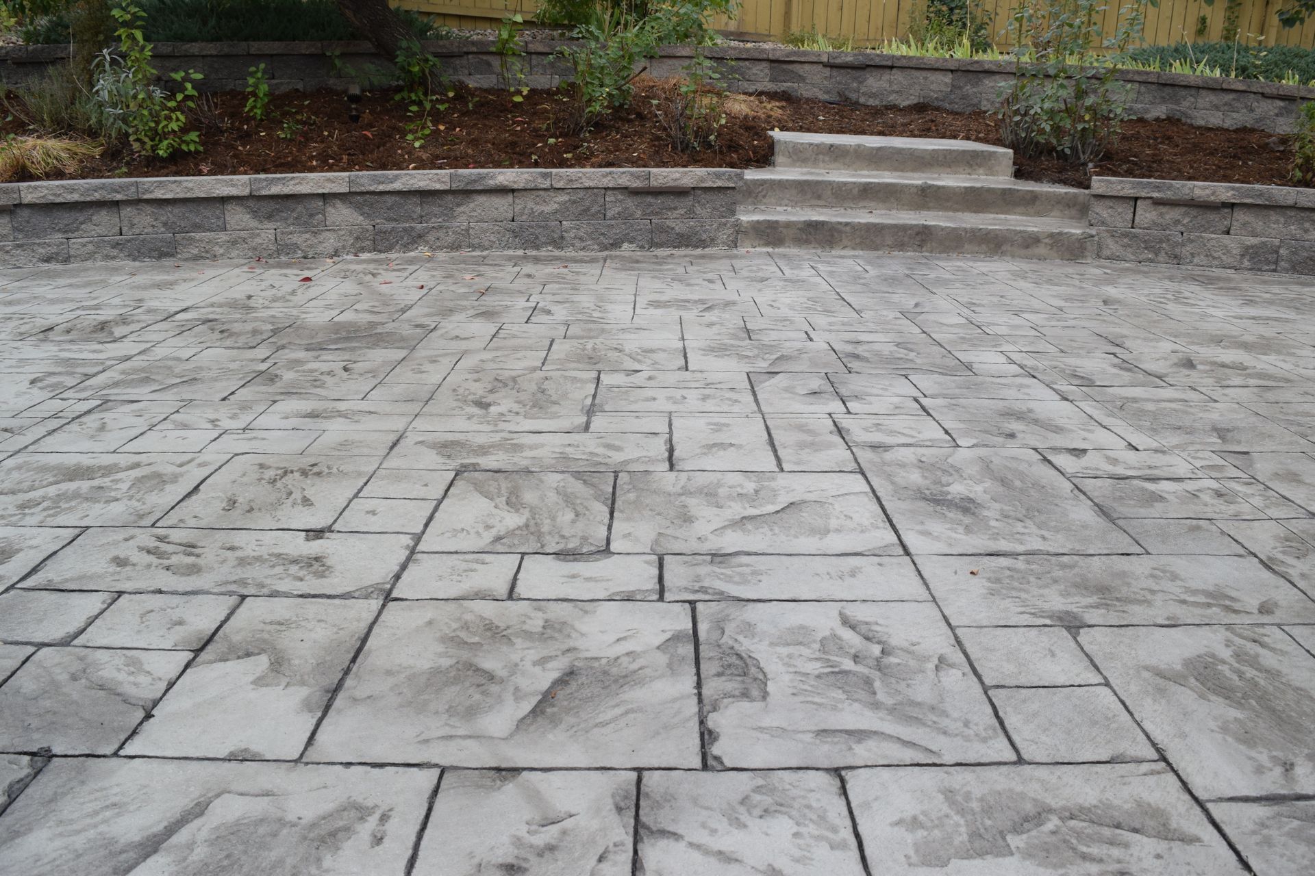 Gray stamped concrete patio with stone pattern, leading up to steps and a raised garden bed.