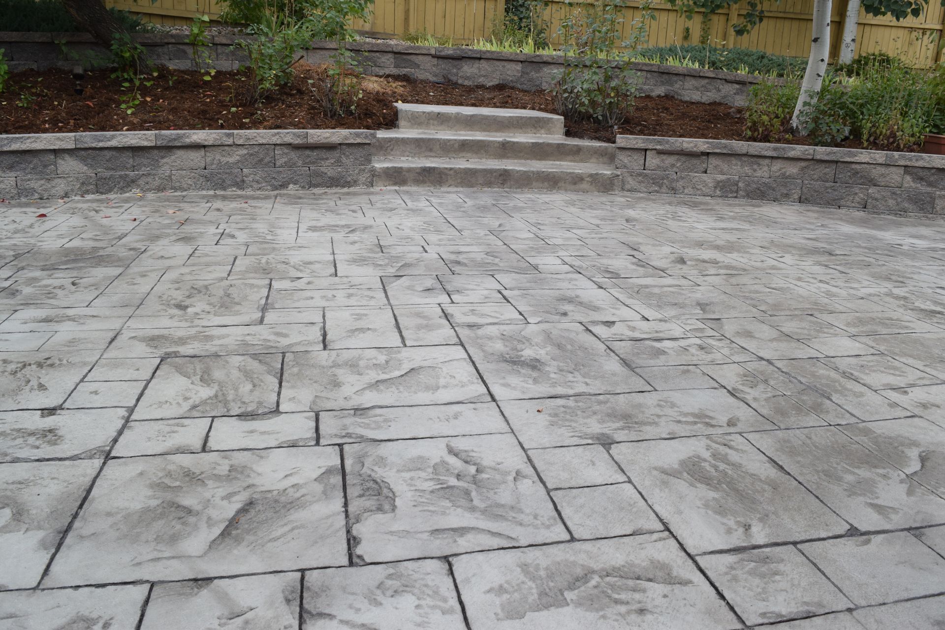 Concrete patio with stone pattern, steps leading to retaining wall, and green foliage.