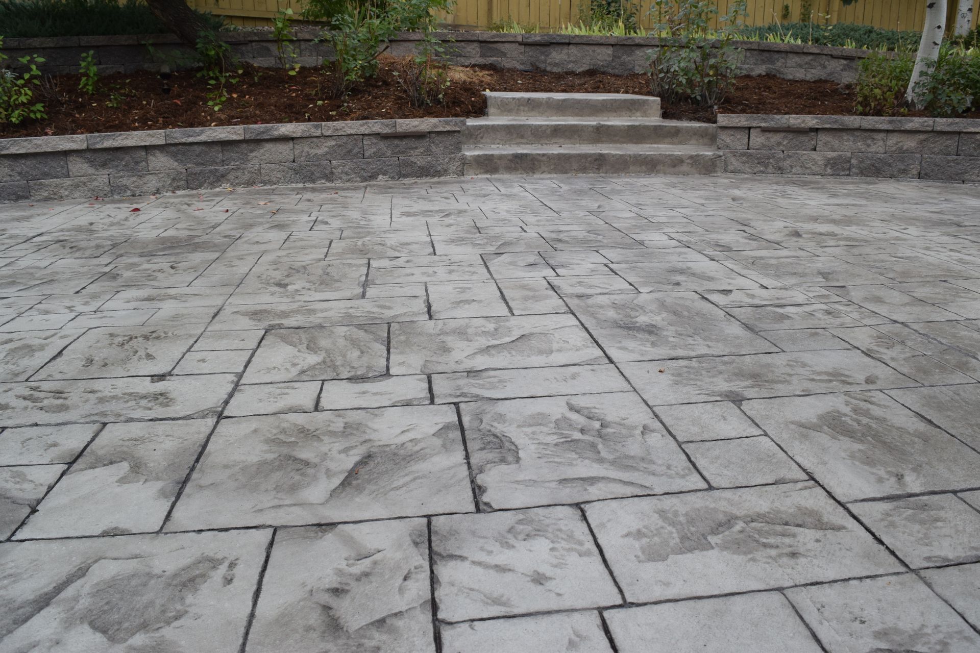 Concrete patio with stone pattern, steps leading to a raised garden bed with plants.
