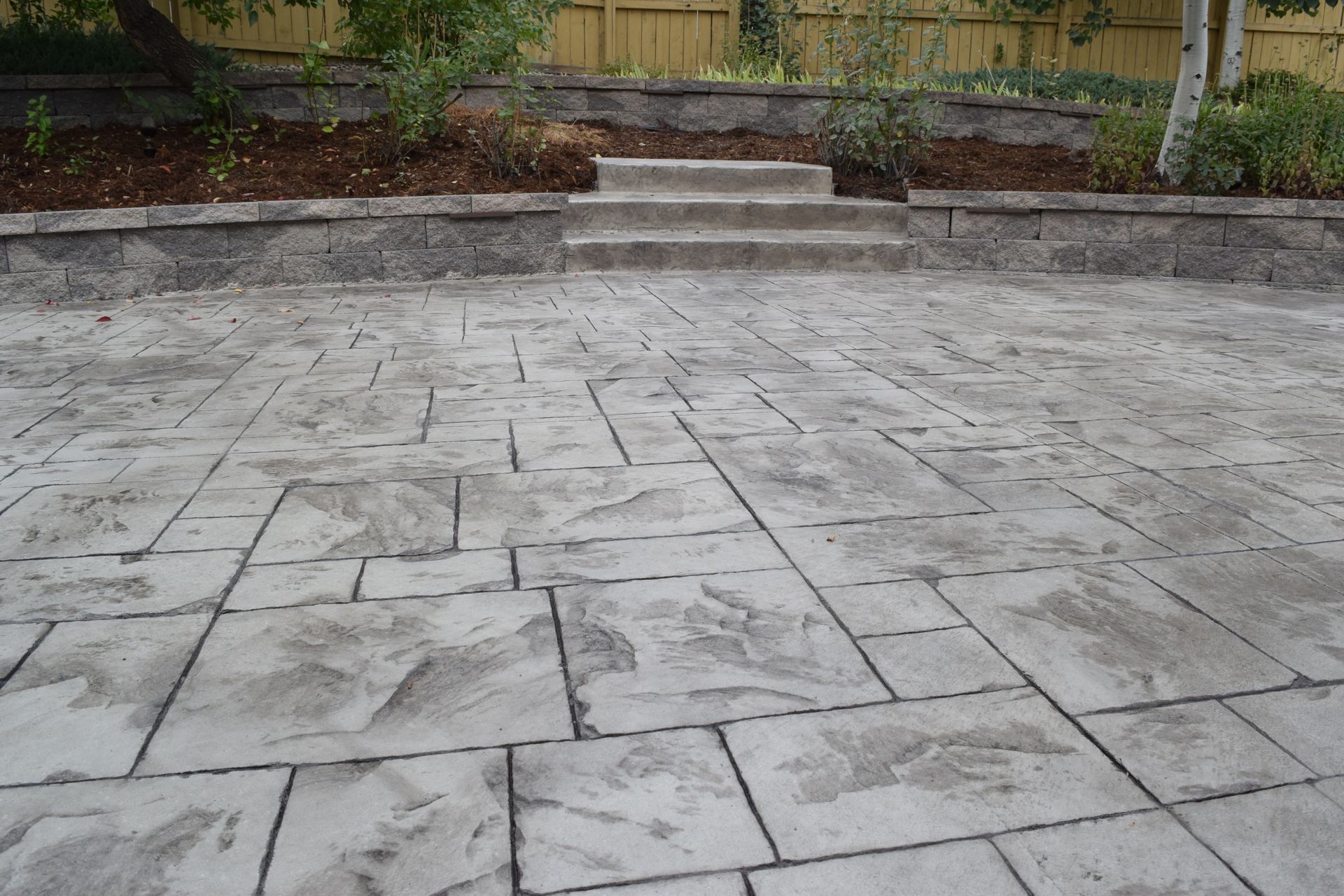 Stone patio with concrete steps leading to a raised garden bed with a wooden fence background.