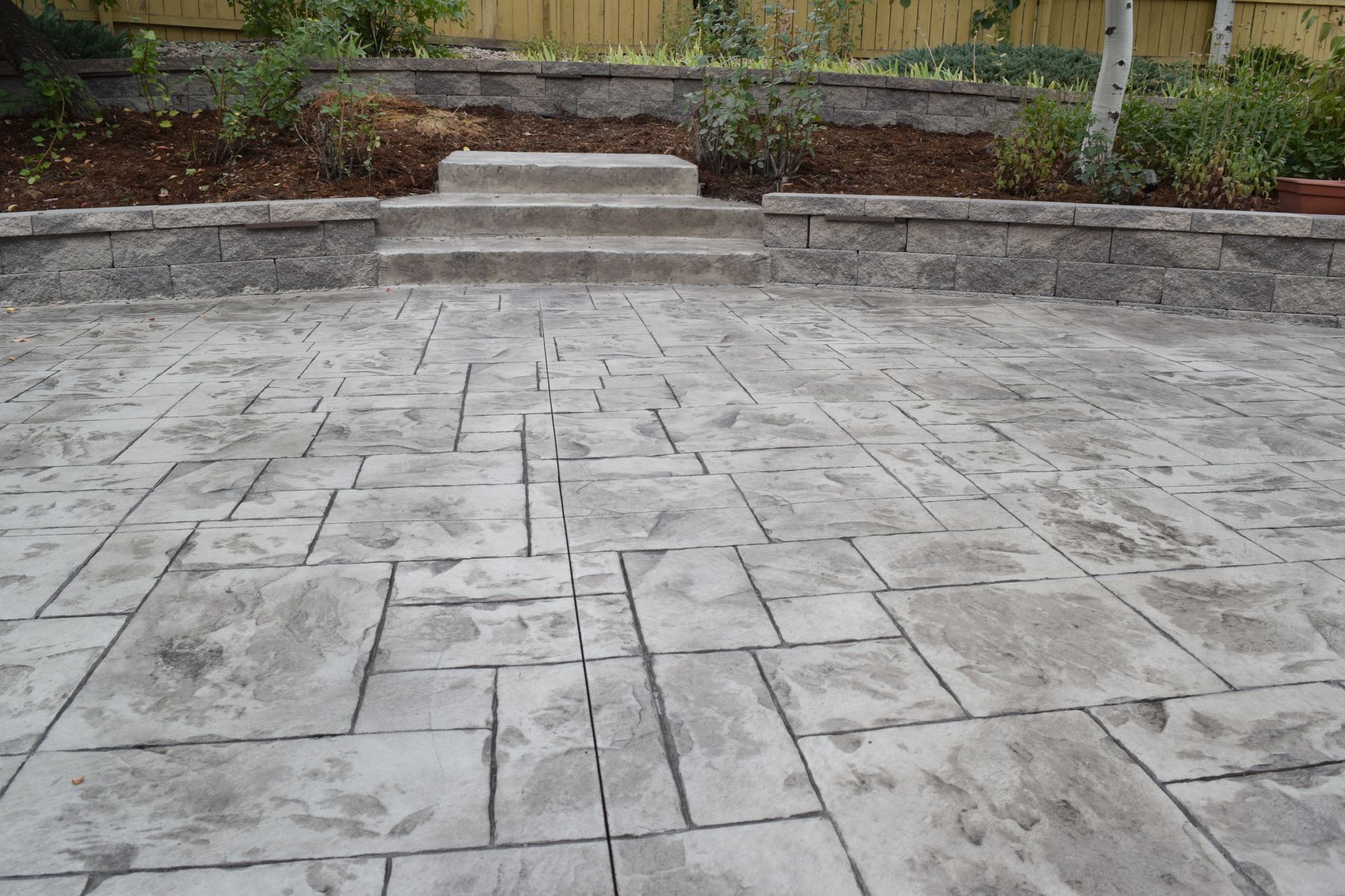 Gray, stamped concrete patio with stone steps leading to a raised garden bed with plants.