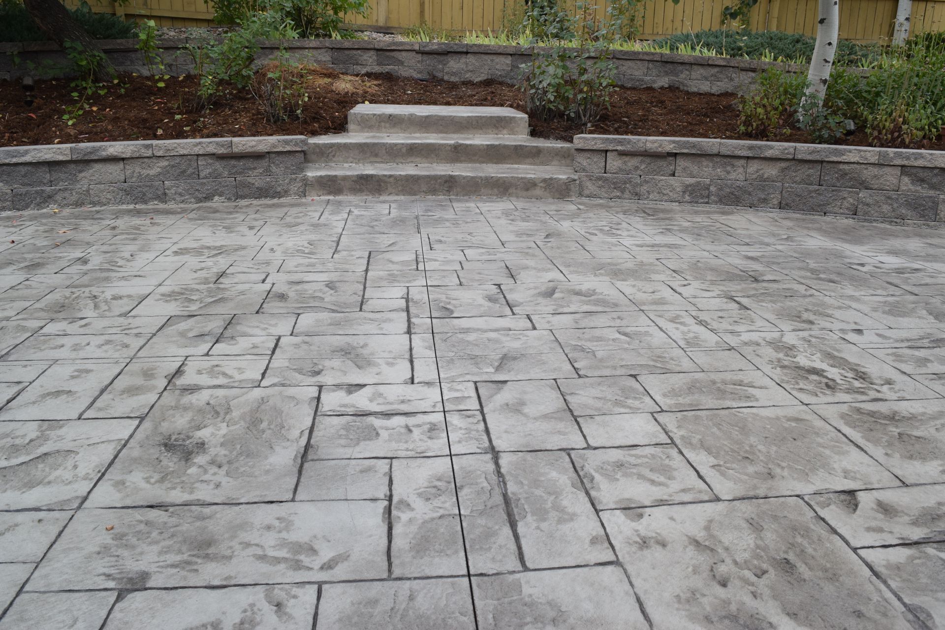 Grey concrete patio with stamped rectangular pattern, leading to steps and a raised garden bed.