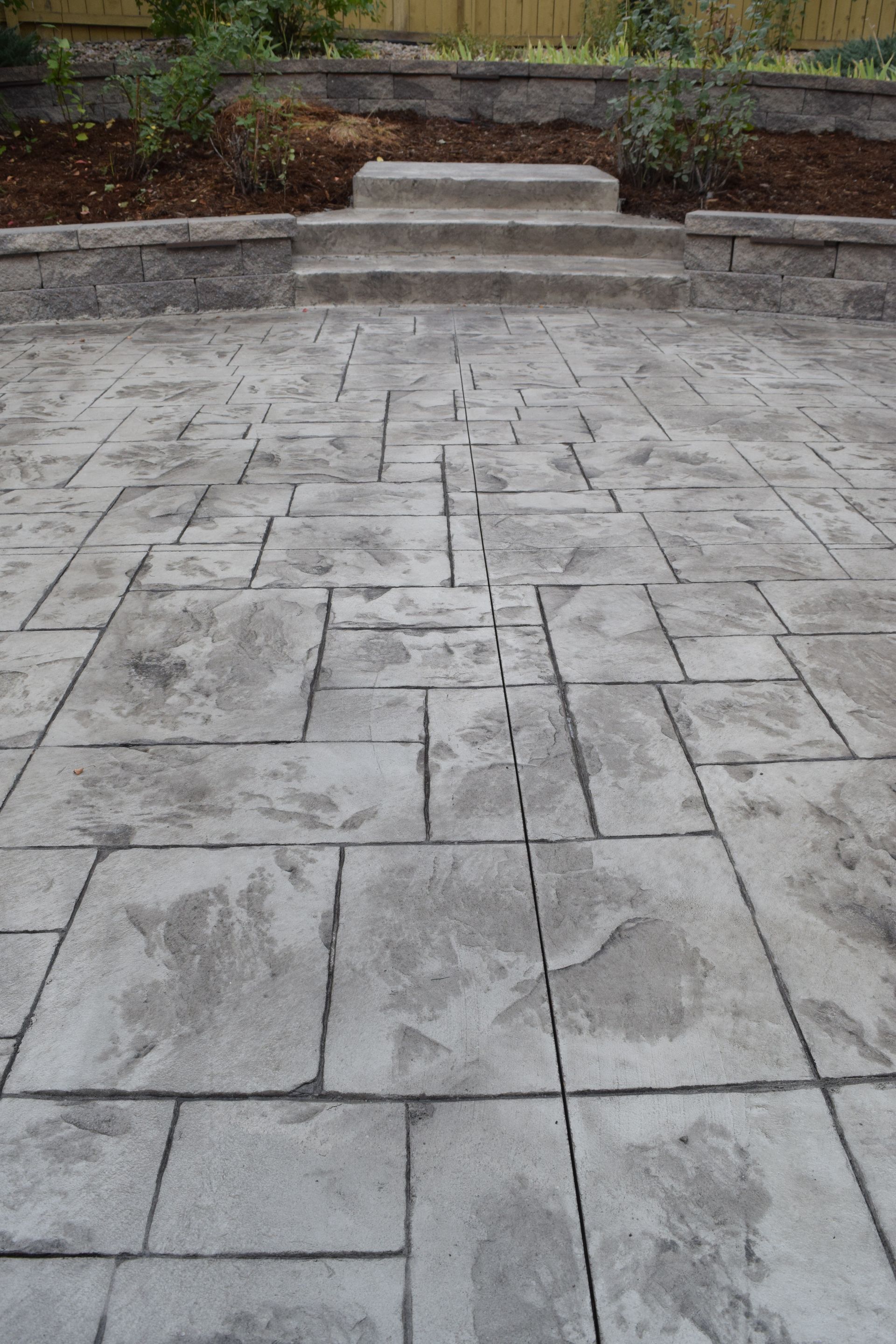 Gray stamped concrete patio leading to stone steps and a retaining wall, with a garden in the background.