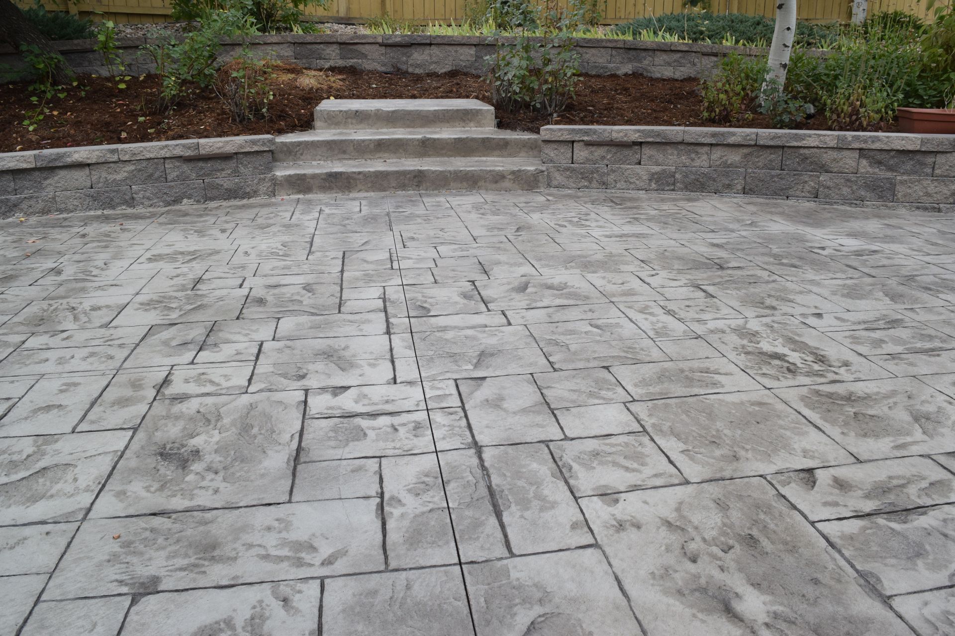 Stamped concrete patio with steps leading to a raised garden bed with plants and a wooden fence.