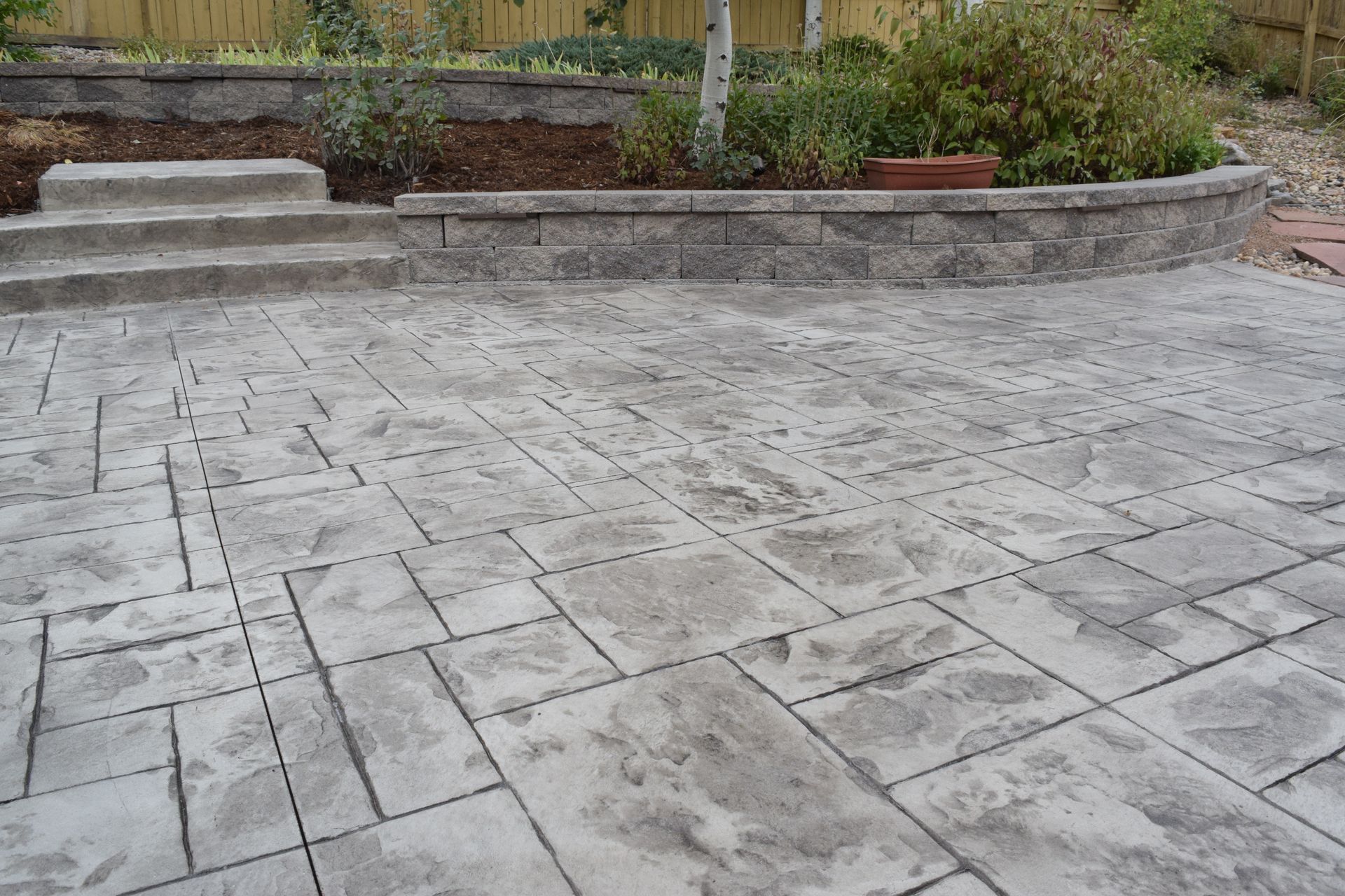 Concrete patio with textured pattern, steps leading to a retaining wall with plants.