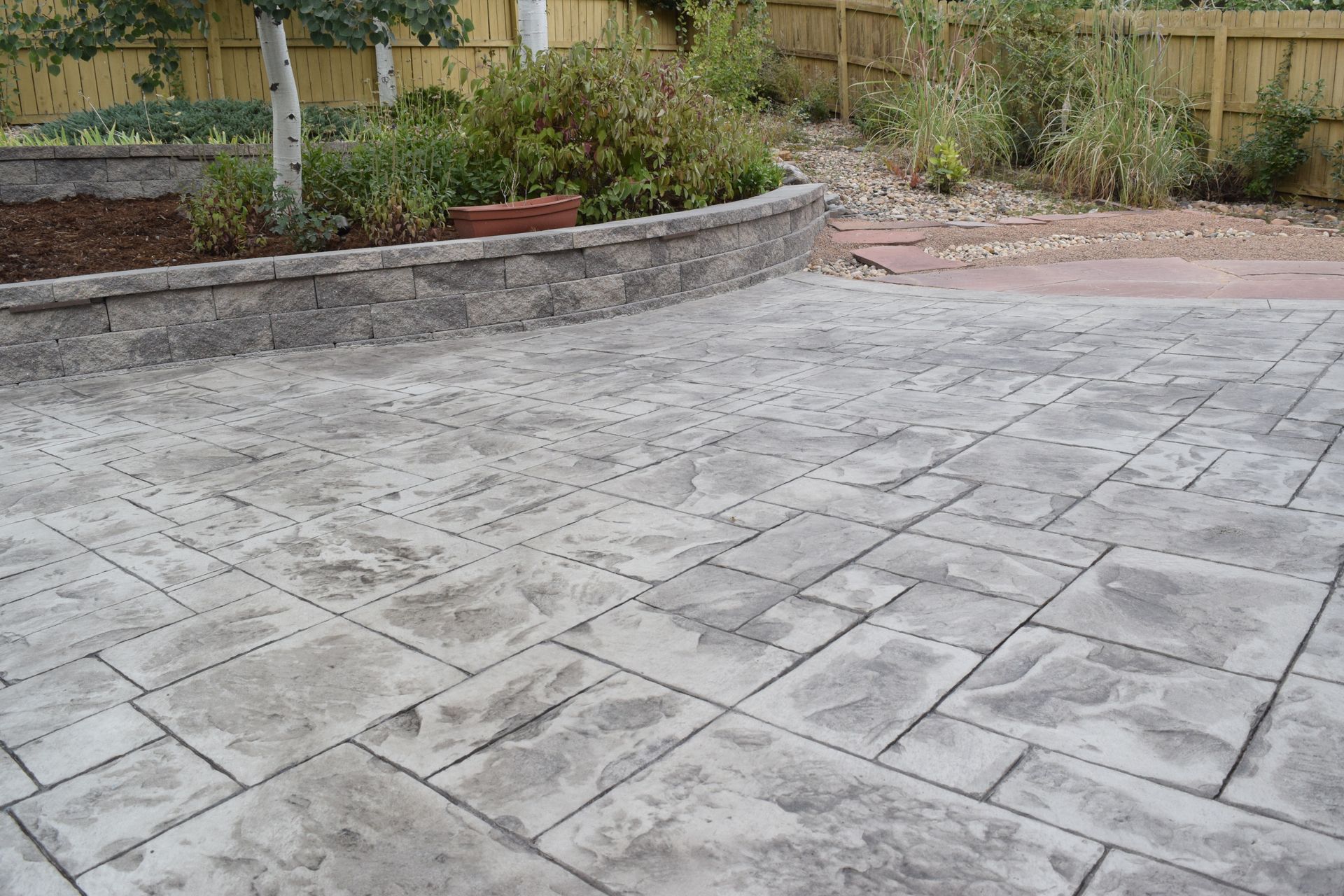 Stamped concrete patio with a curved retaining wall and greenery, in a backyard setting.