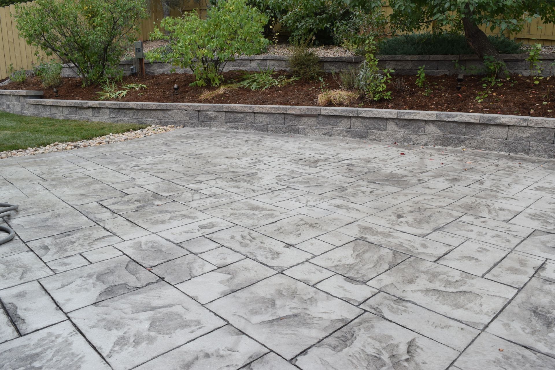 Concrete patio with stone pattern, bordered by a raised garden bed with mulch and shrubs.