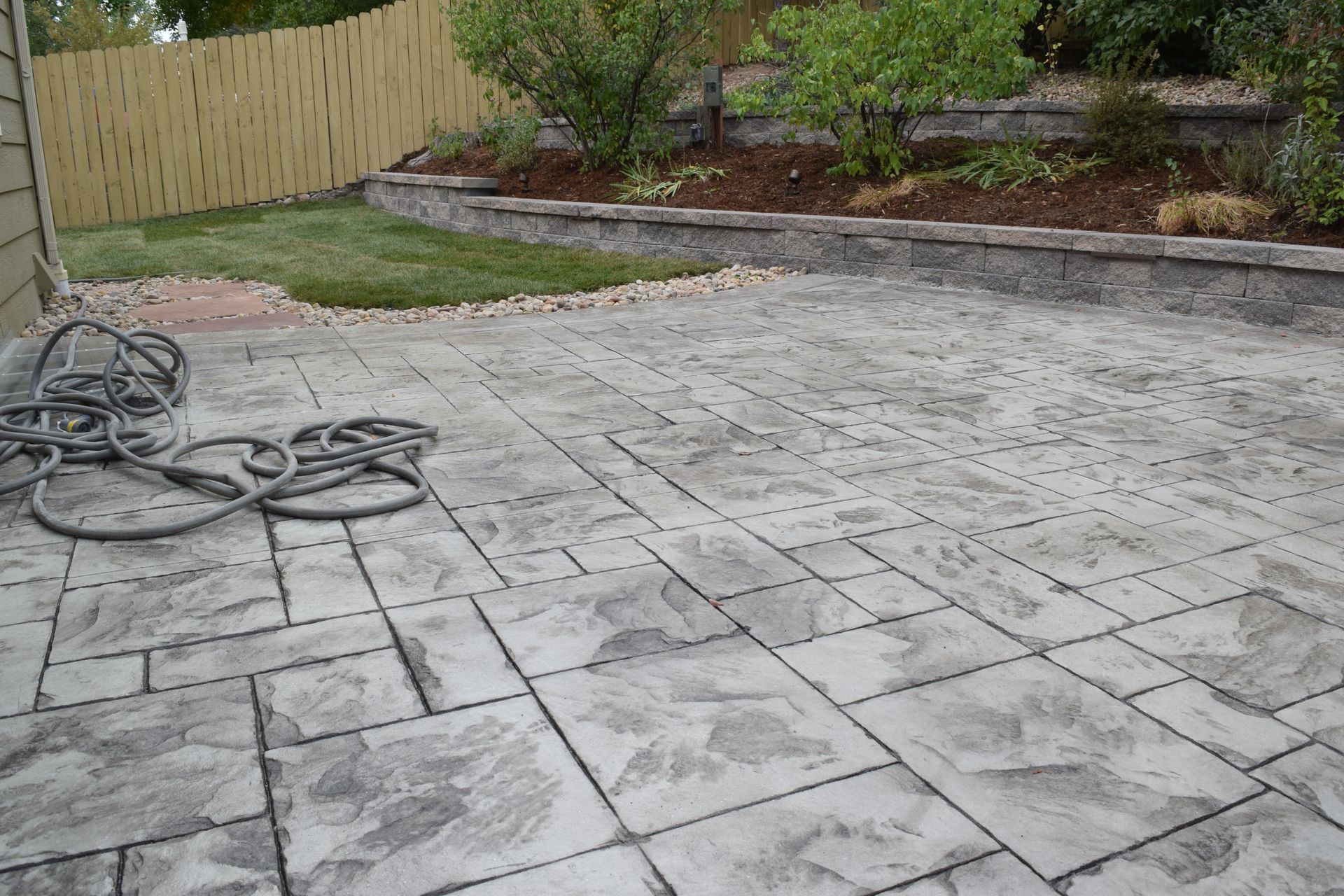 Concrete patio with stone-pattern design. A hose lays on the patio near a small grass area with a fence.