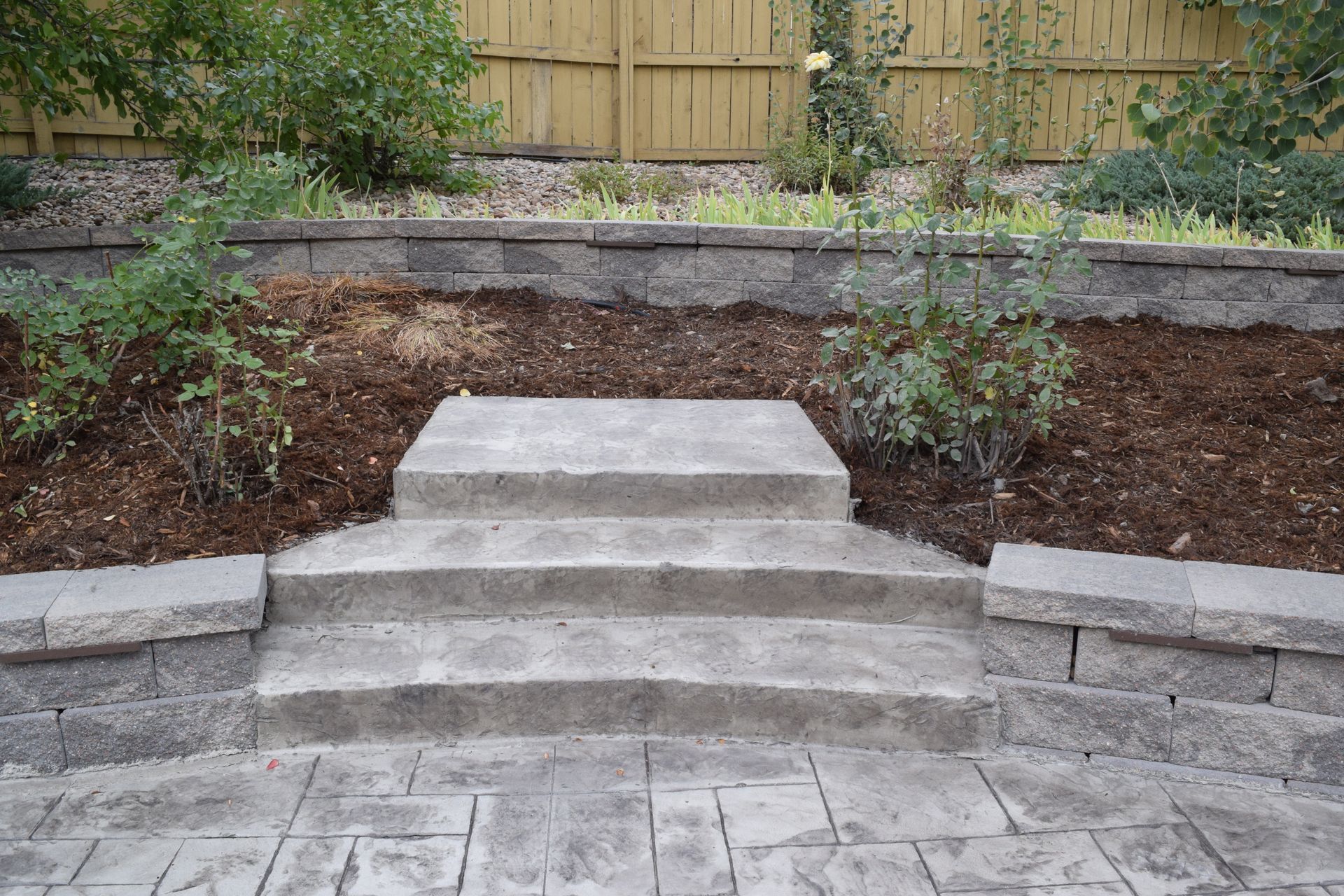 Stone steps leading up to a mulched garden bed with plants, against a wooden fence.