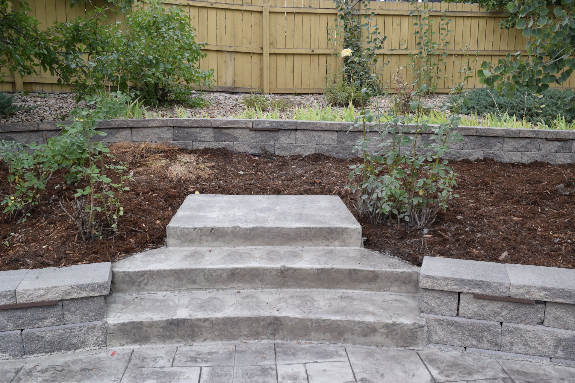 Stone steps leading up to a mulched garden bed with trees and a wooden fence backdrop.