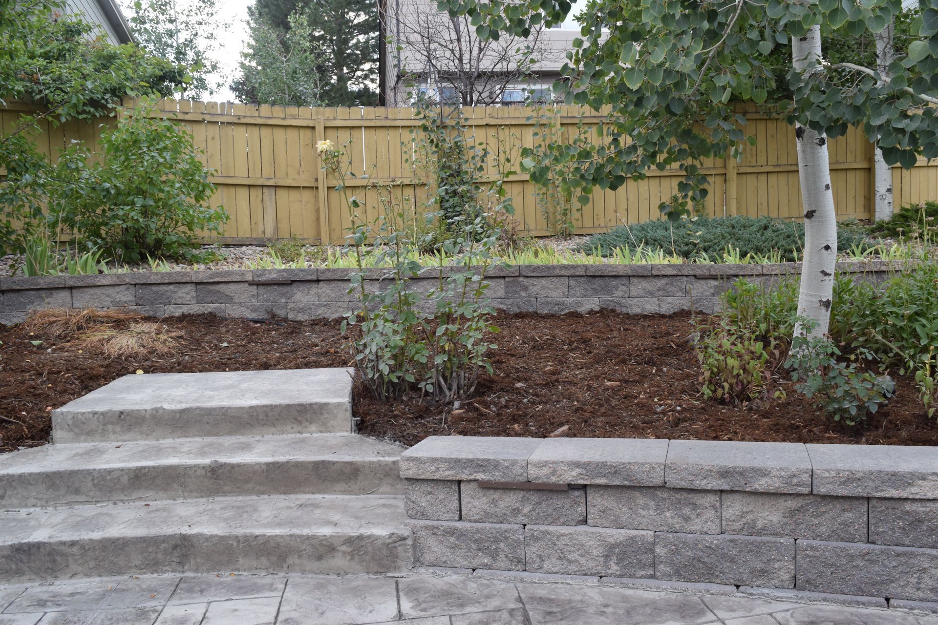 Stone steps and retaining walls in a backyard, with a wooden fence and trees.
