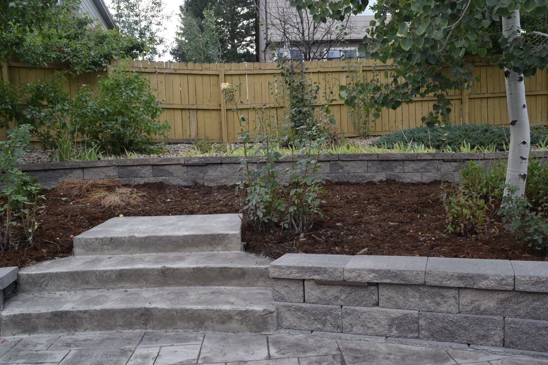 Stone steps lead up to a raised garden bed with mulch and a wooden fence background.
