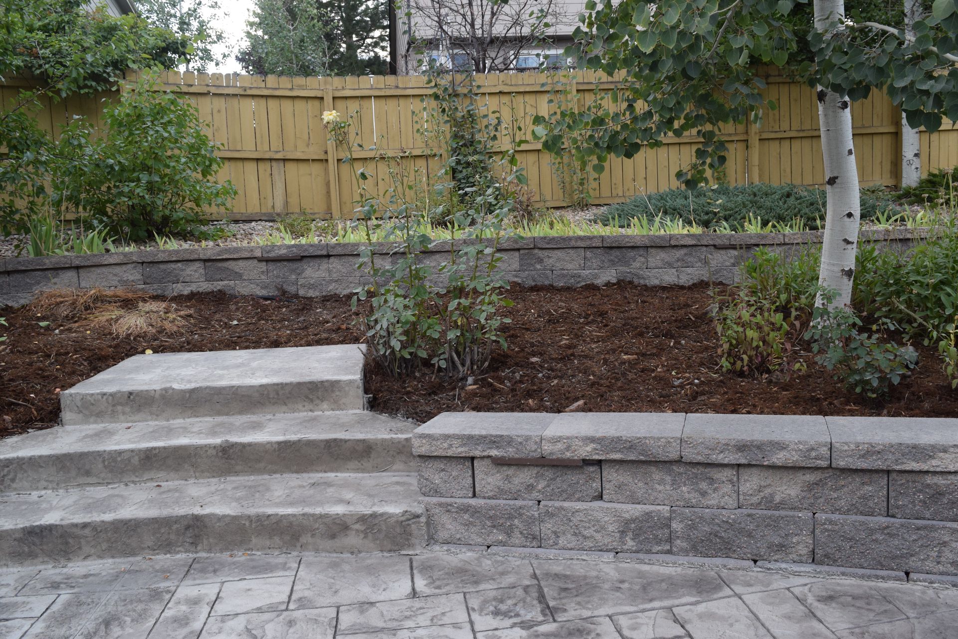 Stone steps and retaining wall in a backyard with trees and a wooden fence.
