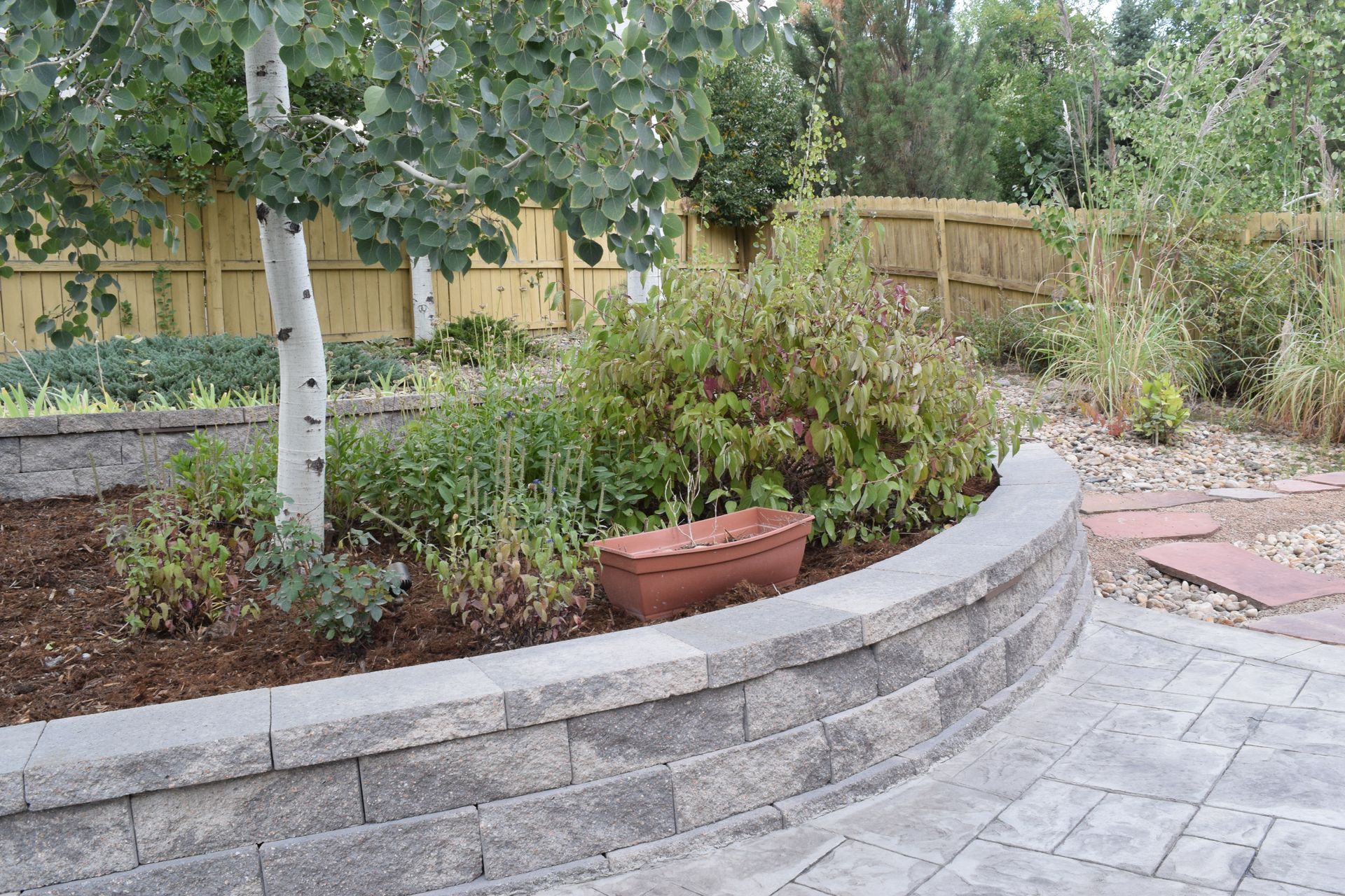 Curved retaining wall with a garden bed, tree, and patio. Brown mulch, stone pavers, and a wooden fence.