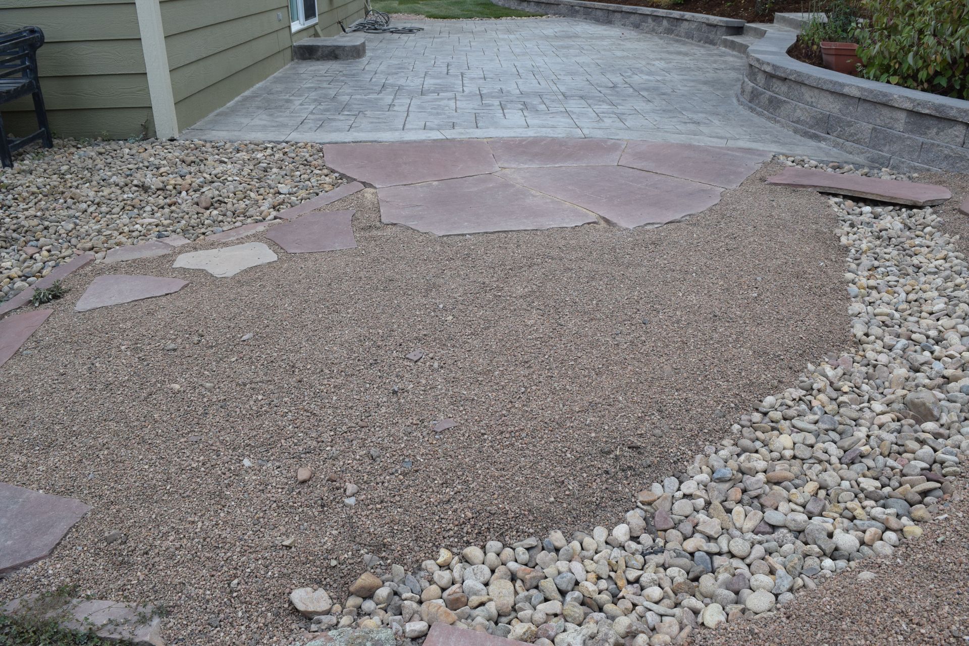 Gravel pathway with stepping stones and a textured concrete patio, transitioning to a retaining wall and building.