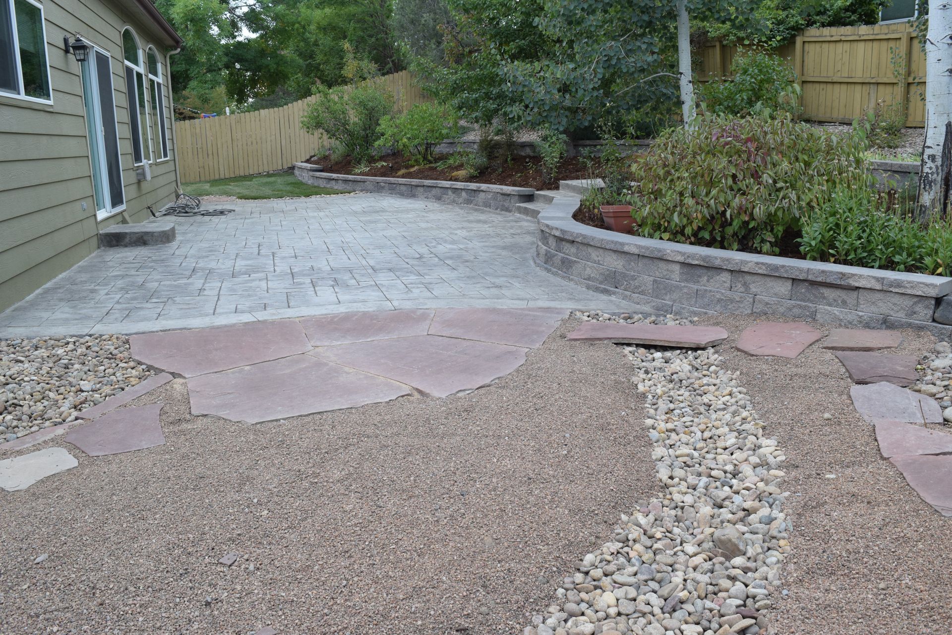 Backyard patio with stamped concrete, gravel path, and retaining wall with plants.
