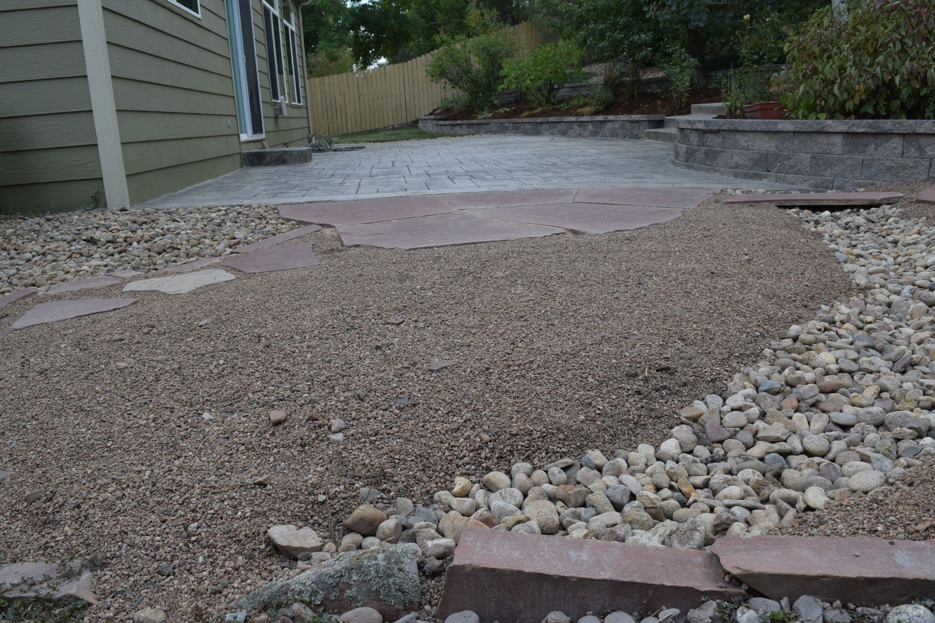 Gravel and stone path leads to a concrete patio, next to a building and greenery.