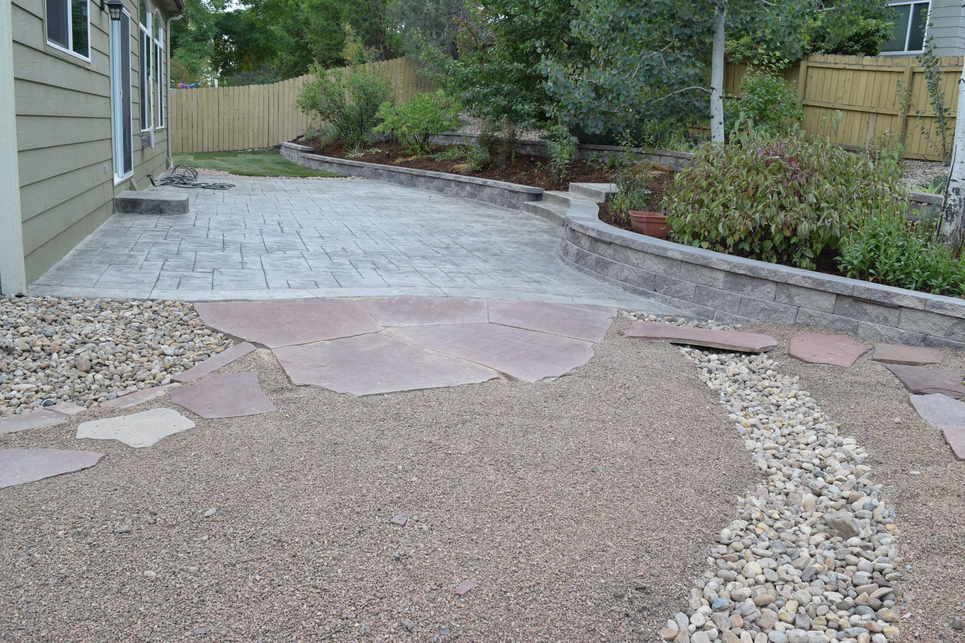 Backyard patio with stamped concrete, gravel path, and retaining wall with plants.