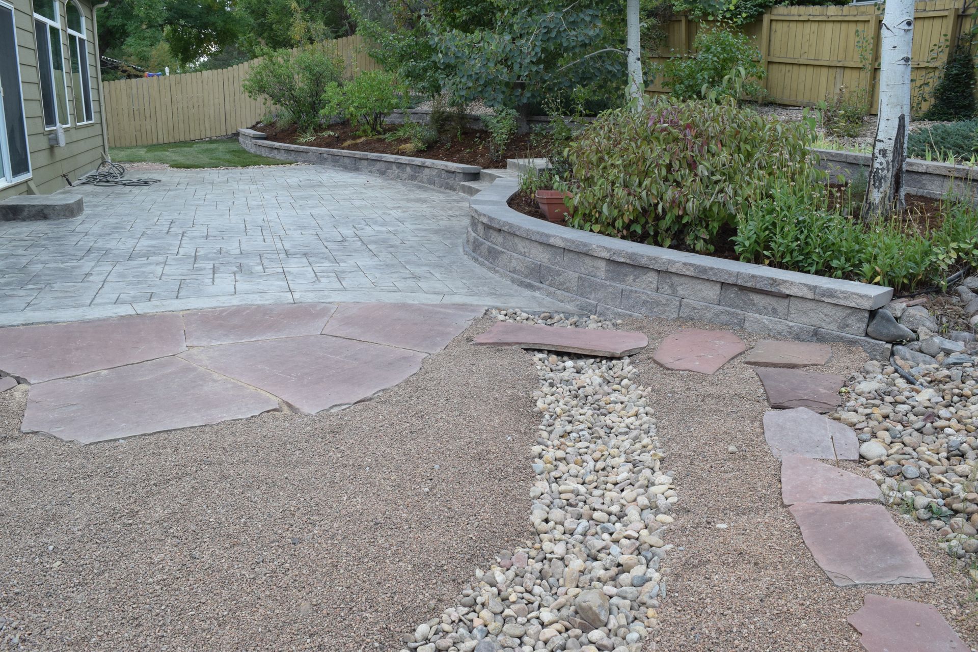 Stone patio with gravel path and retaining wall with shrubs.