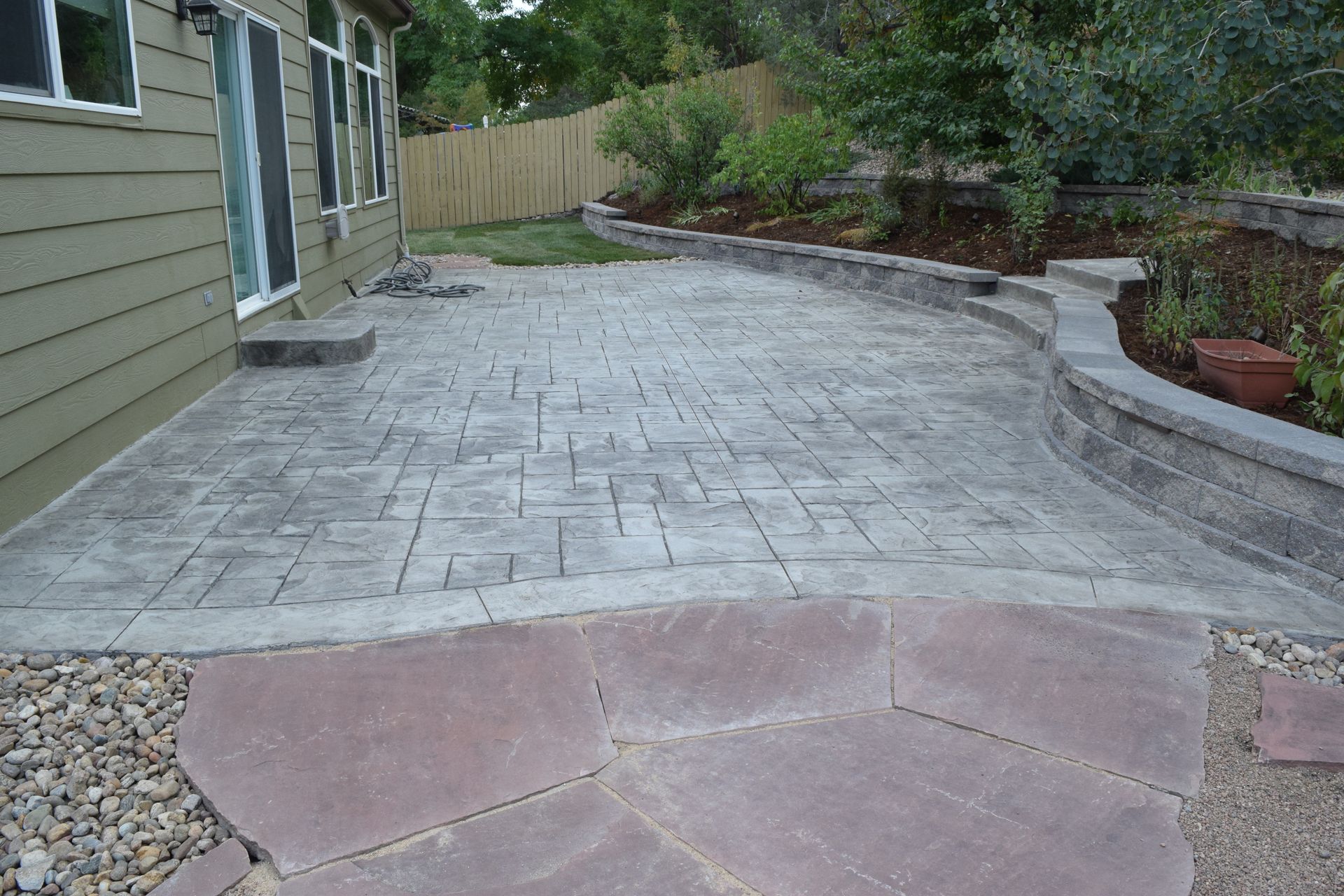 Concrete patio with textured surface, bordered by stone walkway and retaining wall.