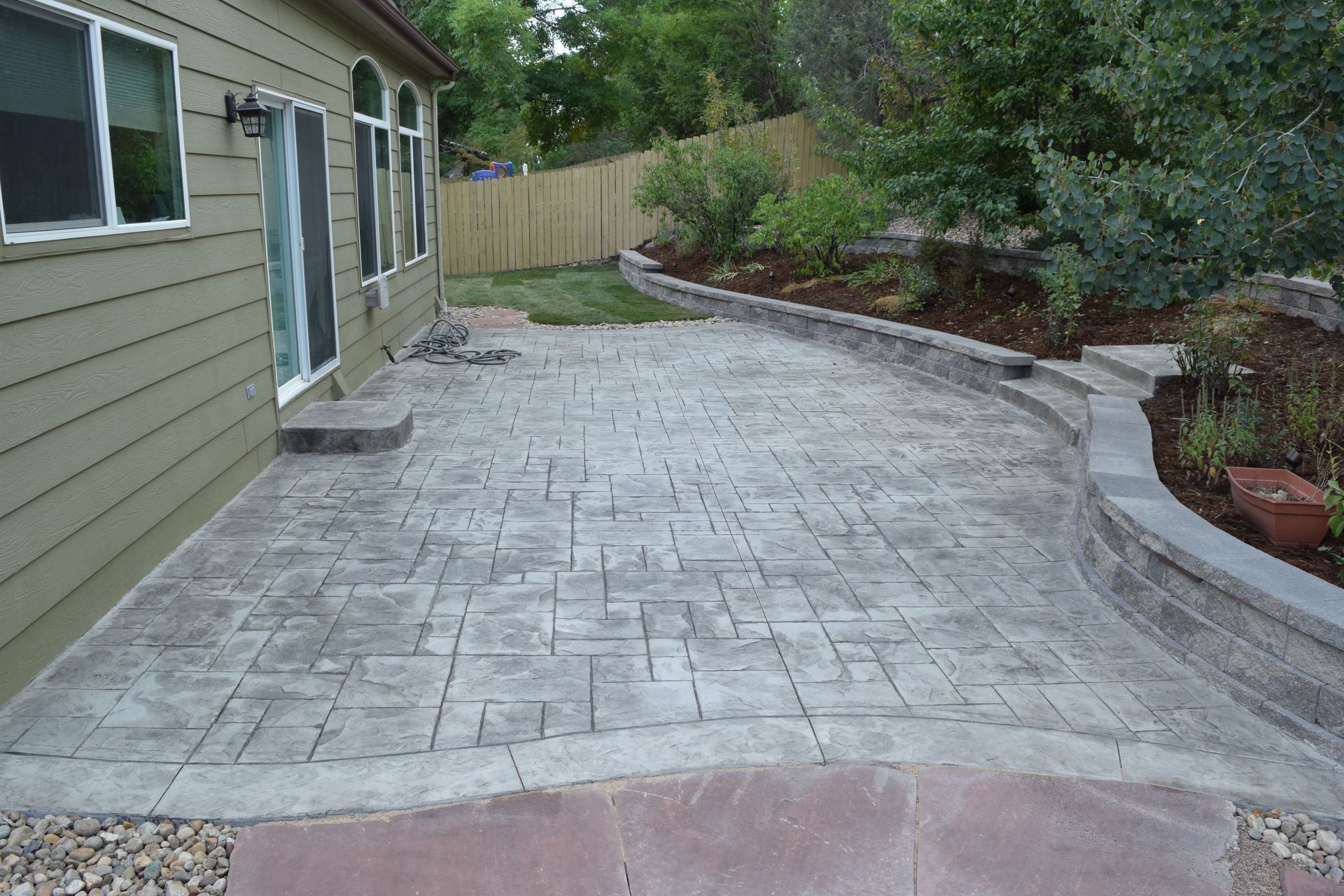 Concrete patio with textured surface, next to a house with a garden bed and fence.