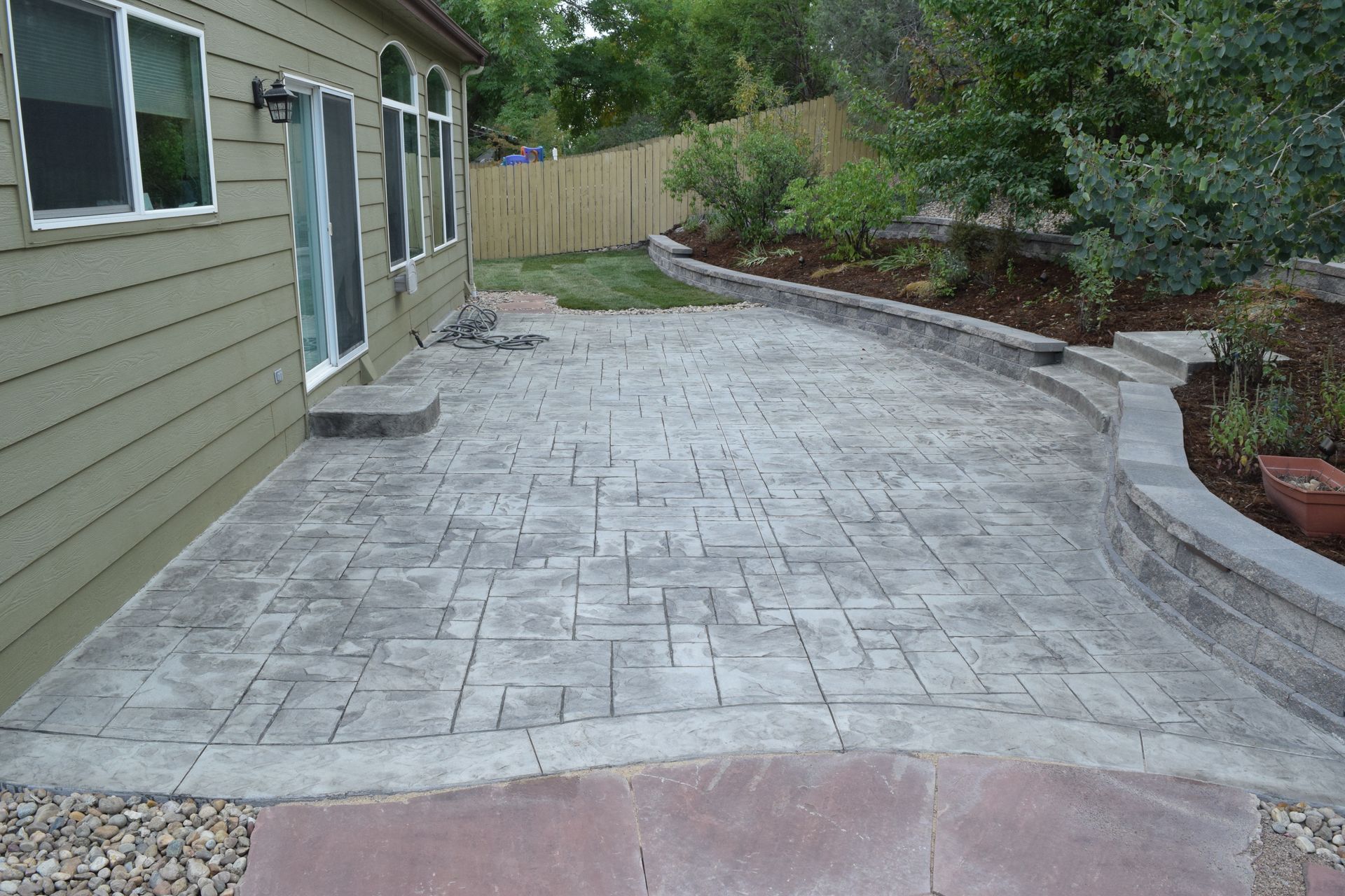 Gray stamped concrete patio next to a house with a retaining wall and plants.