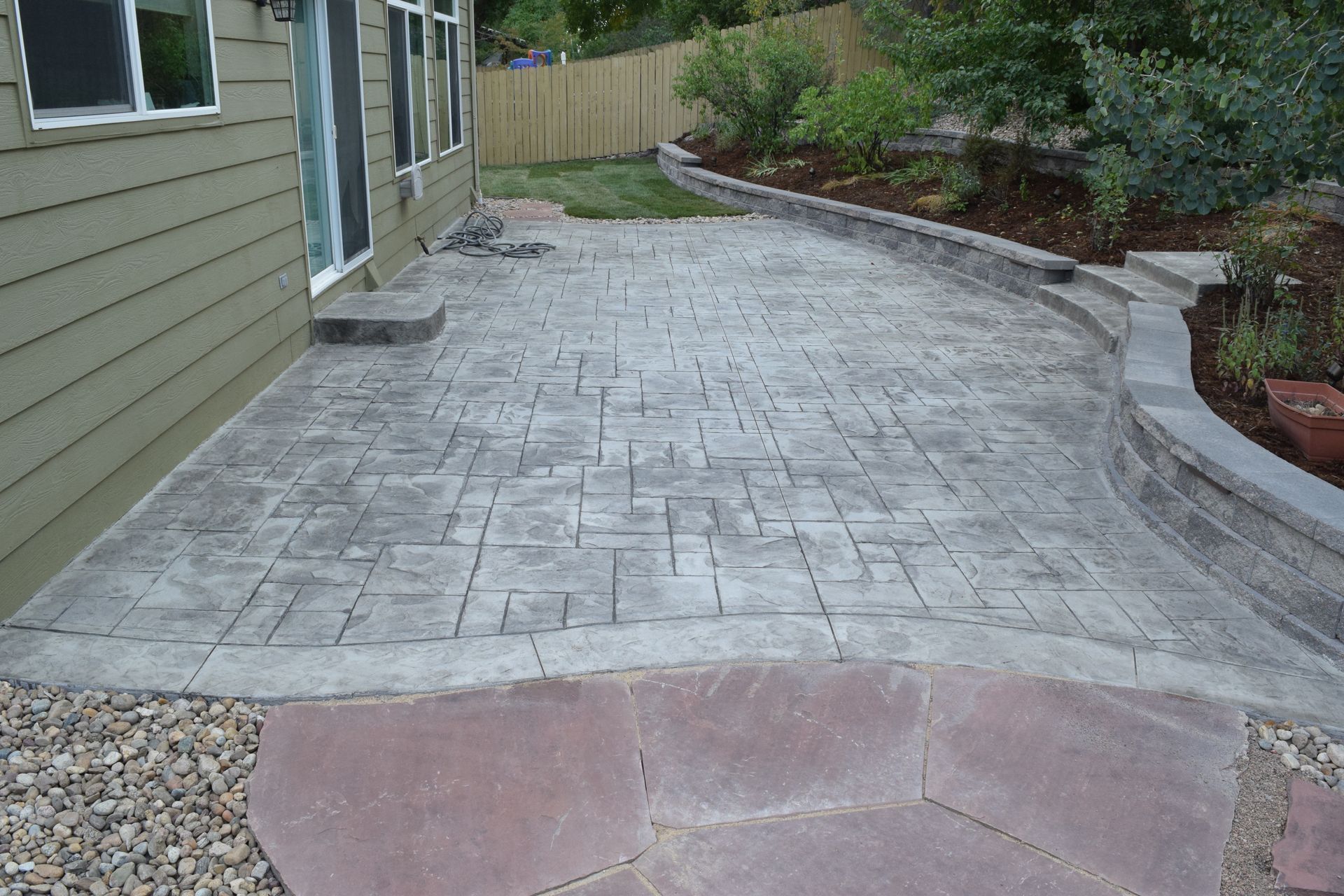 Concrete patio with stamped pattern, stone border, and retaining wall next to a house with a green exterior.