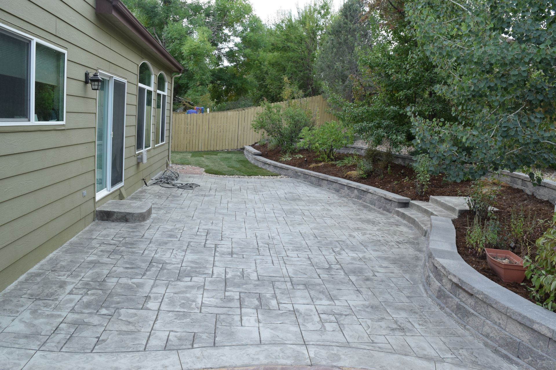 Patio with patterned concrete, bordering a landscaped garden near a house with windows.