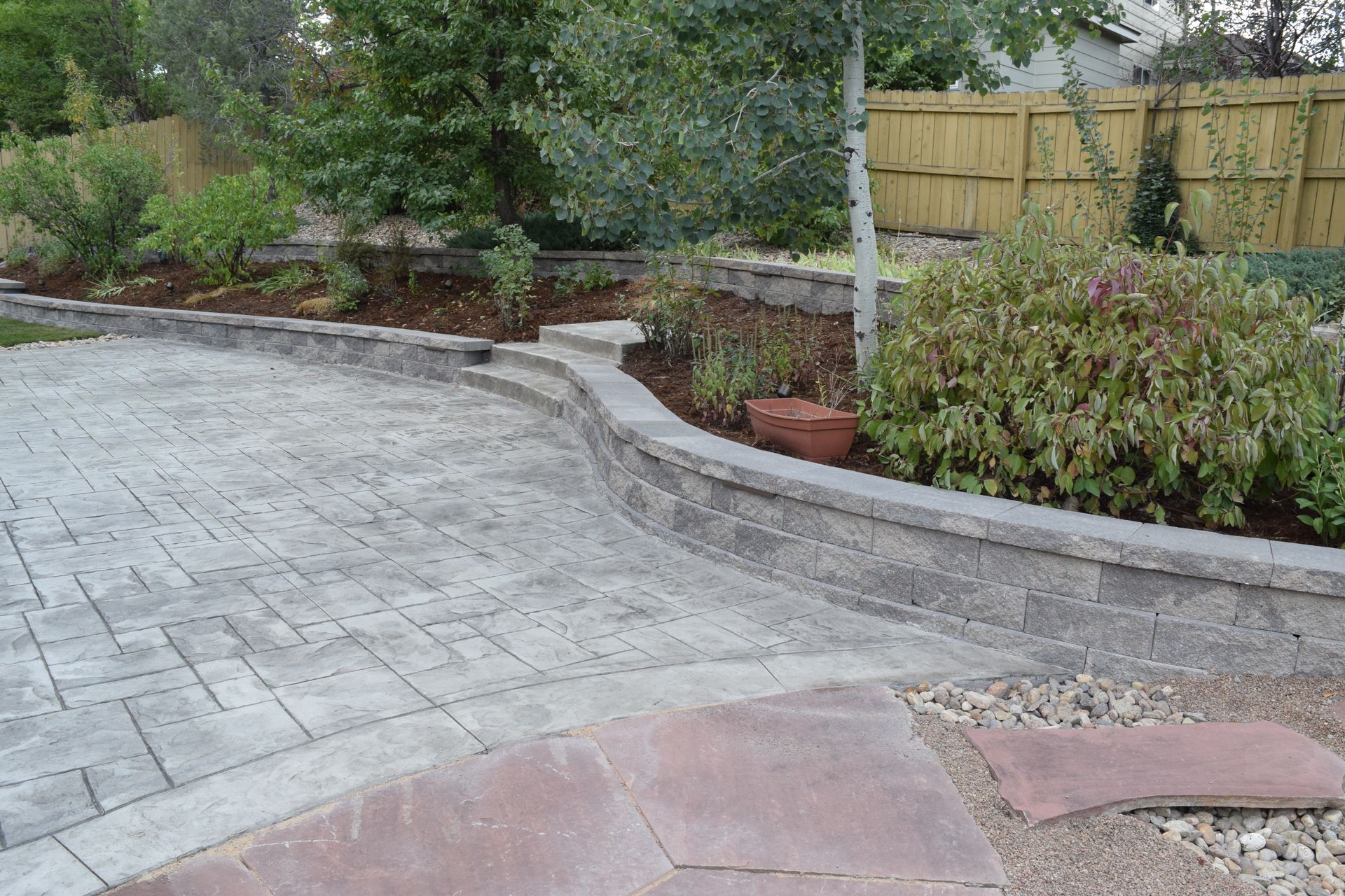 Brick patio with a retaining wall separating a garden bed, against a wooden fence.