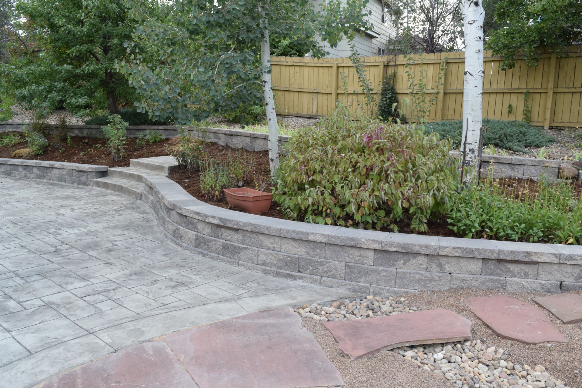 Stone patio curves to meet a landscaped garden with raised beds and trees.