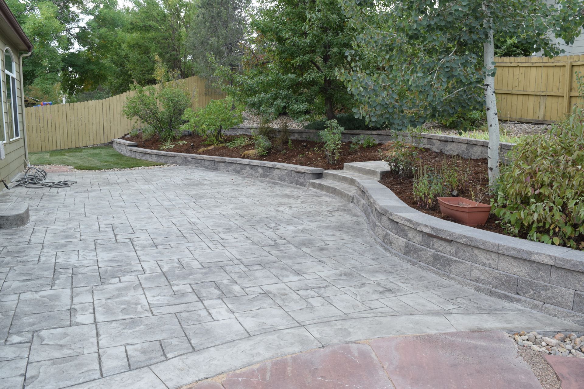 A backyard patio with stamped concrete, retaining wall, and landscaping next to a house with a wooden fence.