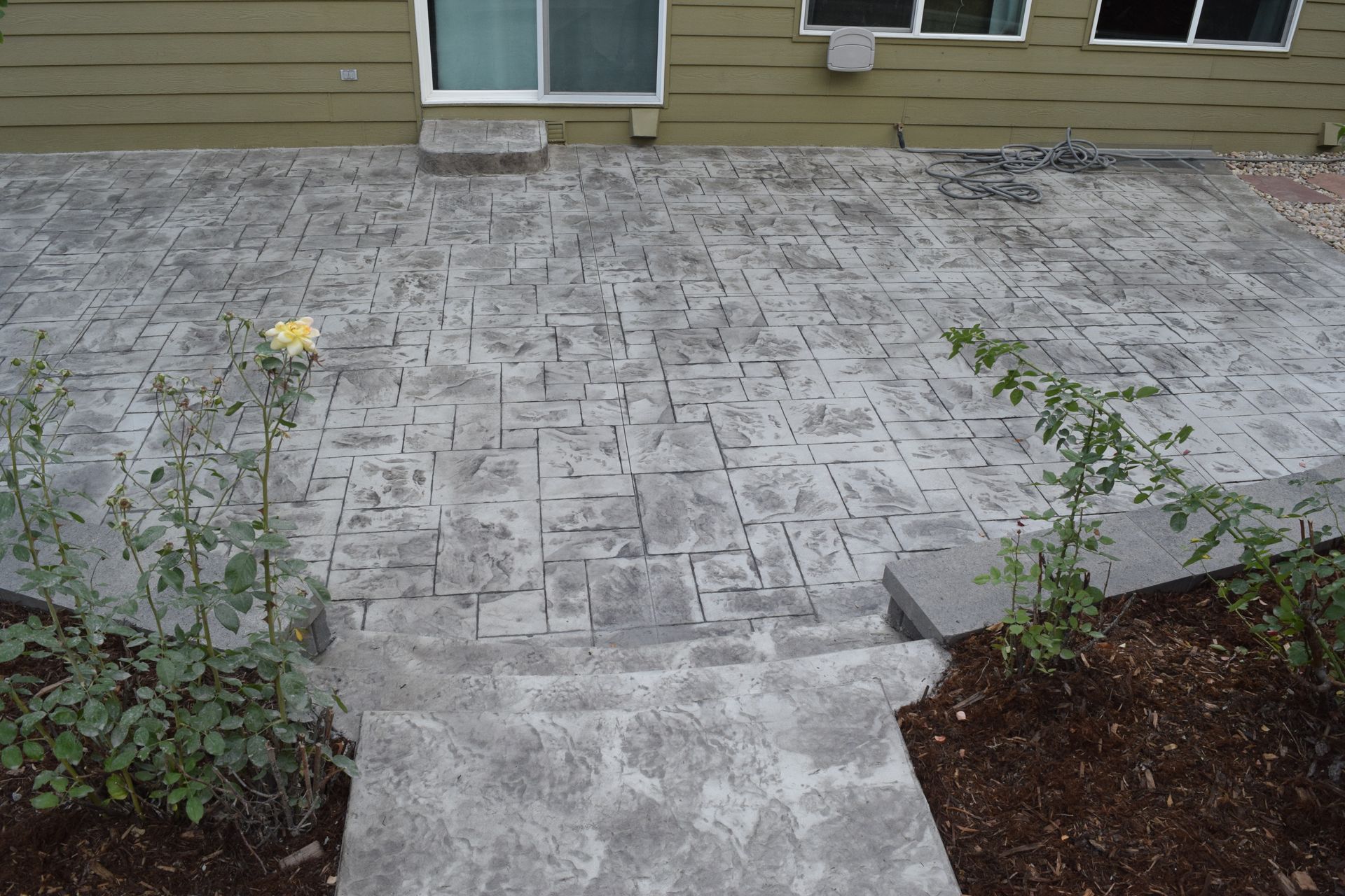 Concrete patio with textured surface and steps leading down to a garden bed with plants.
