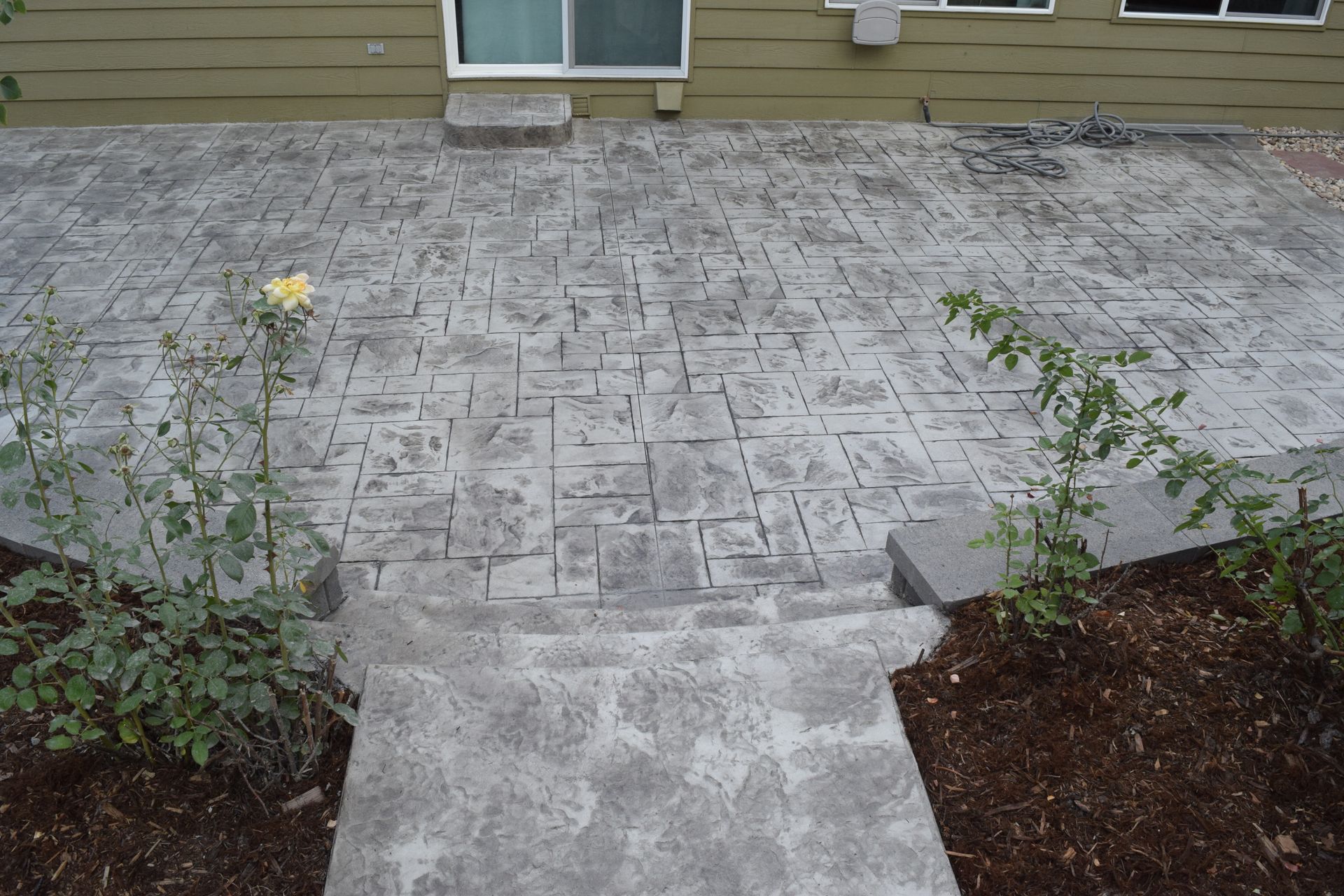 Concrete patio with stamped brick-like pattern, leading to a house with a window, flanked by flower beds.