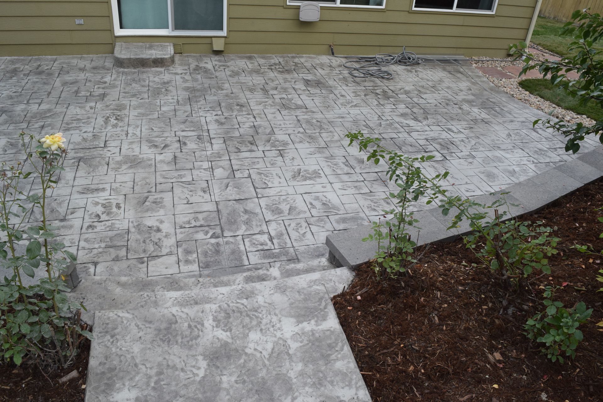 Concrete patio with steps, stamped in a stone pattern next to a house with small plants and mulch.
