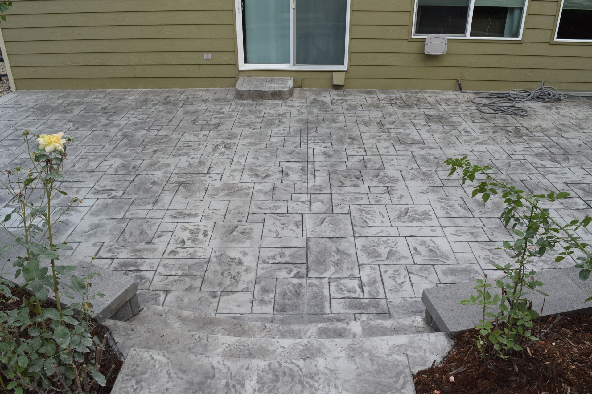 Concrete patio with steps, gray, textured, leading to a house with a sliding glass door and windows, bushes.