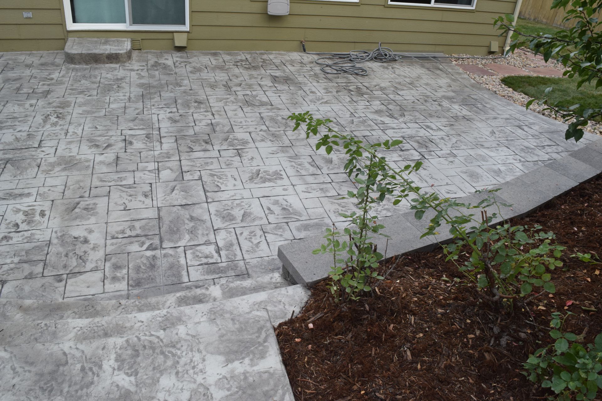 Concrete patio with textured brick pattern and steps, adjacent to a garden with plants and mulch.