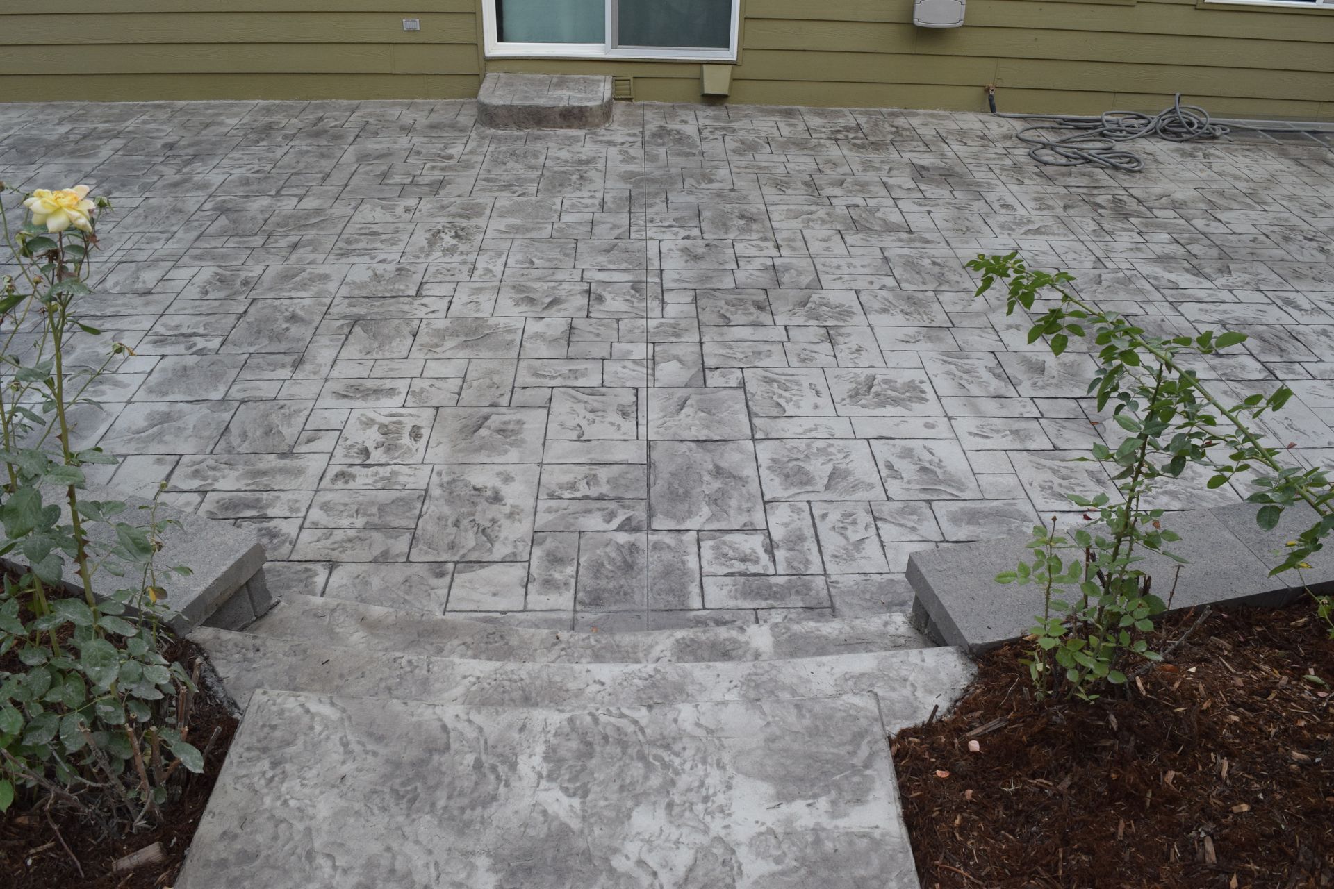 Concrete patio with steps, textured in gray stone pattern, with small rose bushes.