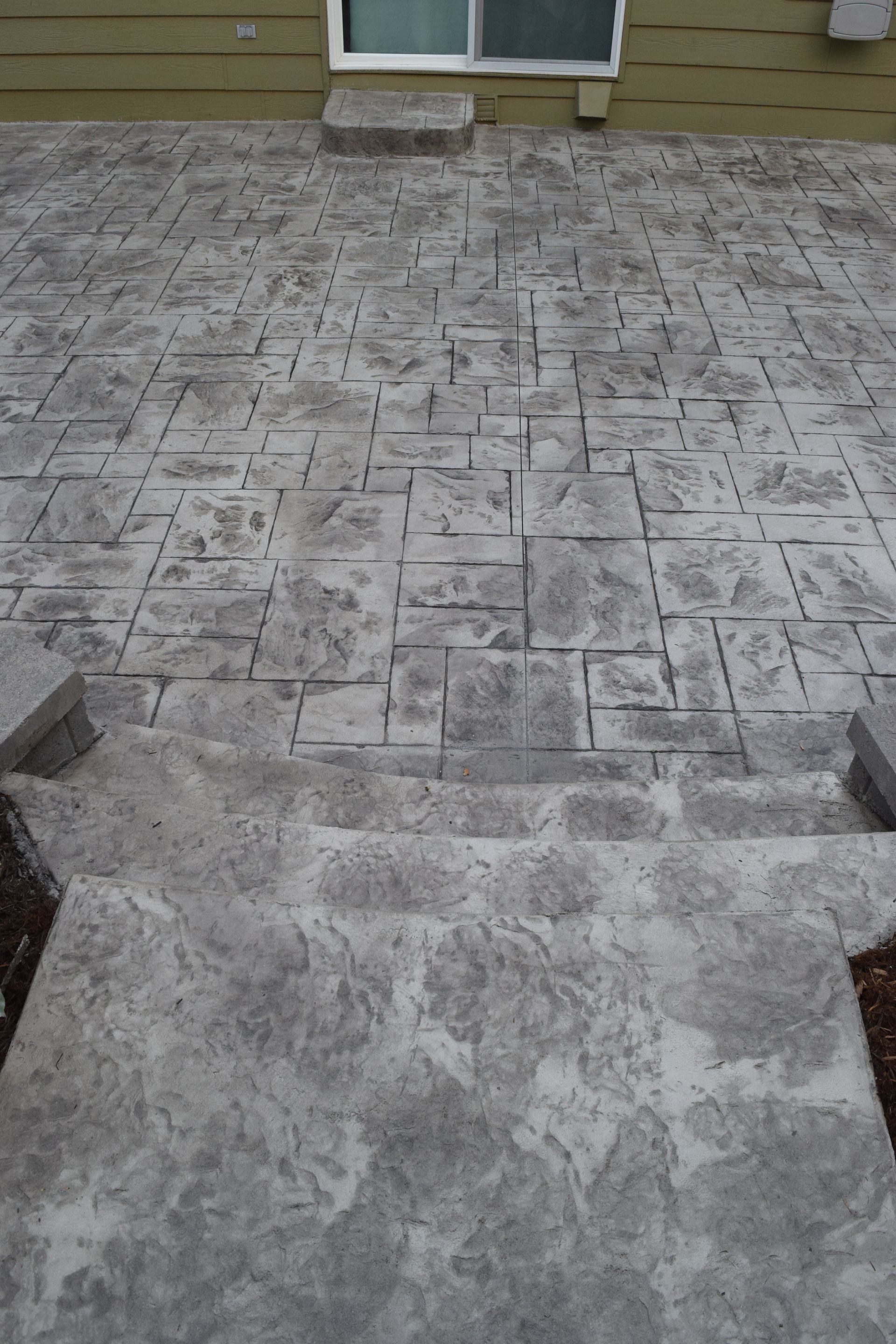 Concrete patio with textured stone pattern, steps leading down from a building with a window.