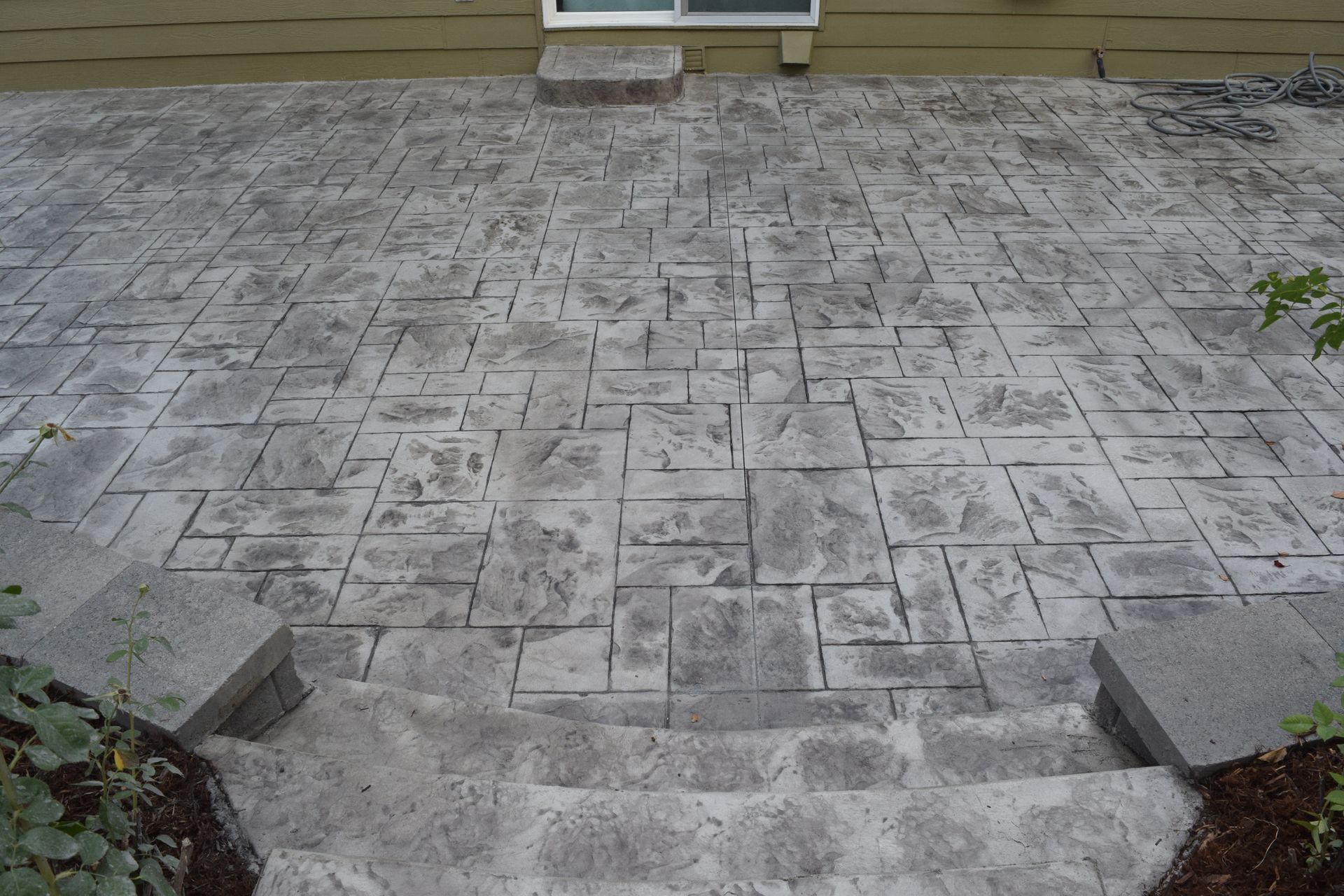 Gray stamped concrete patio with steps leading down from a house with a window.