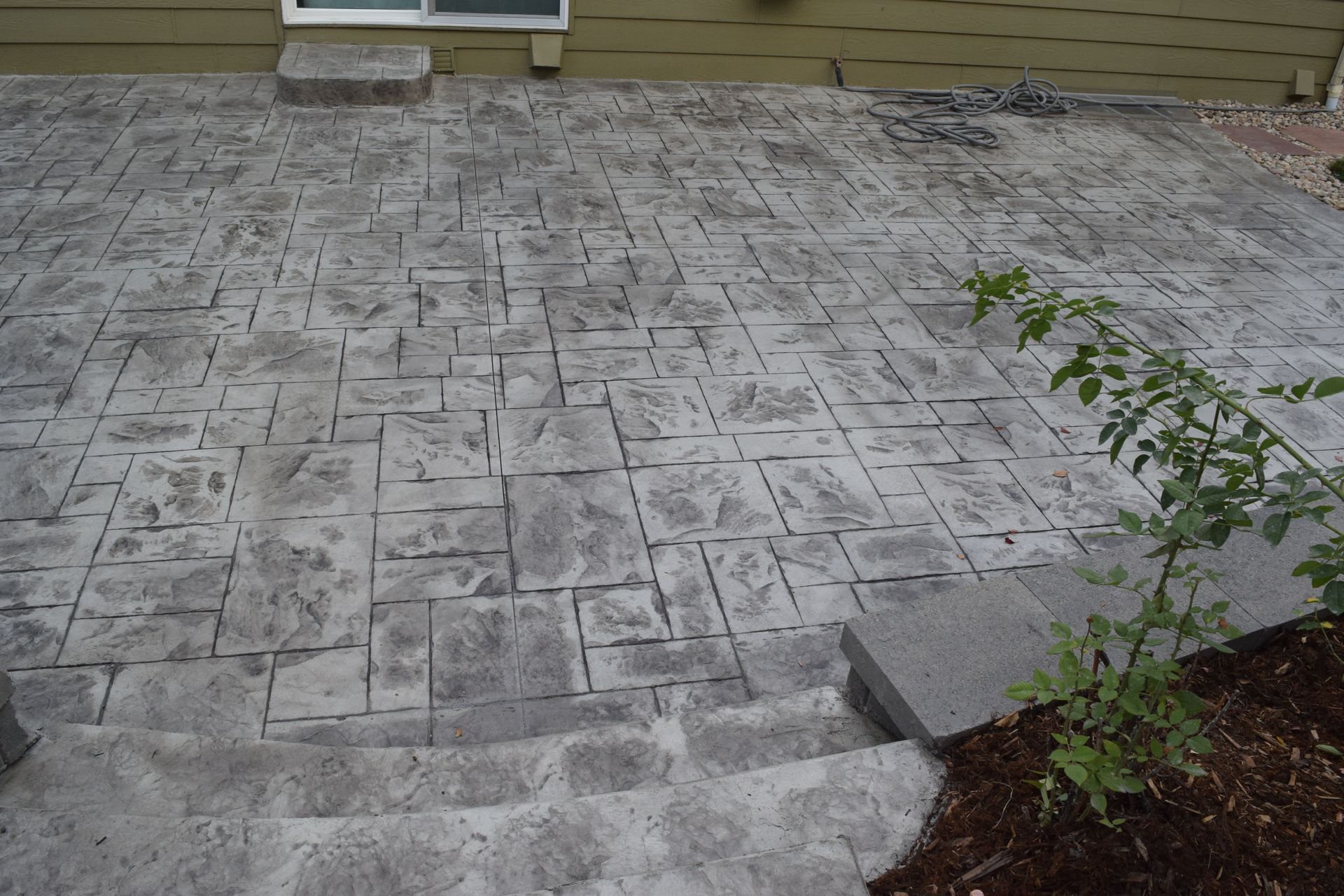 Stamped concrete patio with steps leading down to a garden bed with a green plant, next to a beige house.