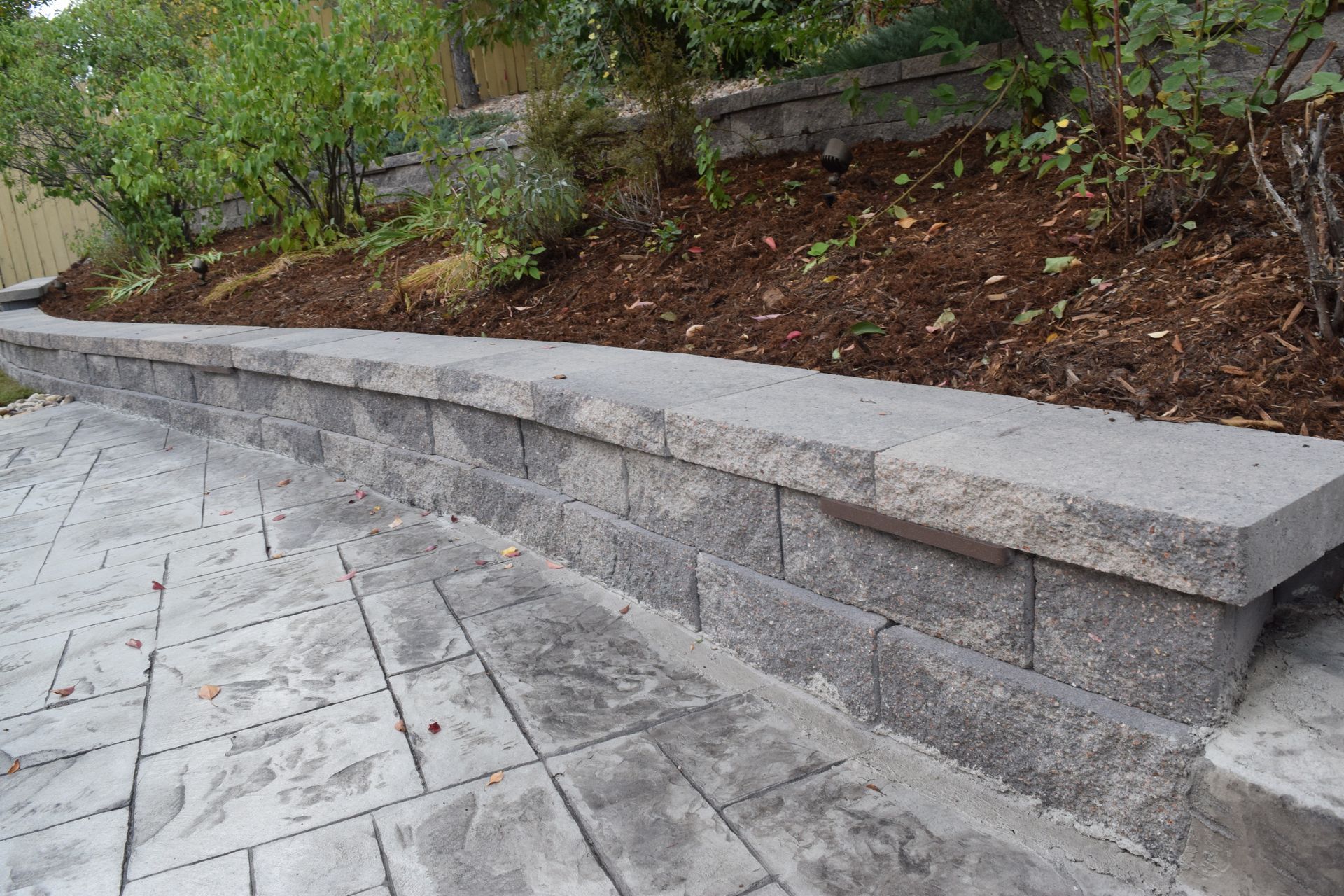 Stone retaining wall with a patio in front; plants and dirt in the background.