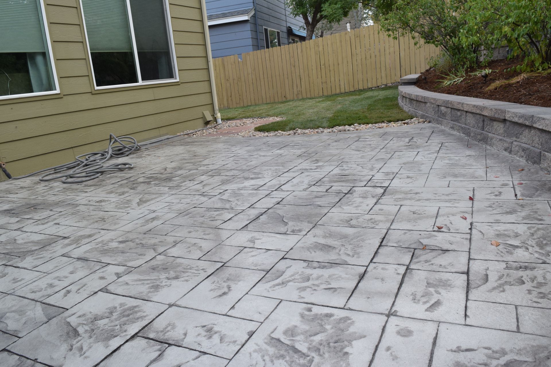 Gray stamped concrete patio next to a house with a retaining wall and fence.