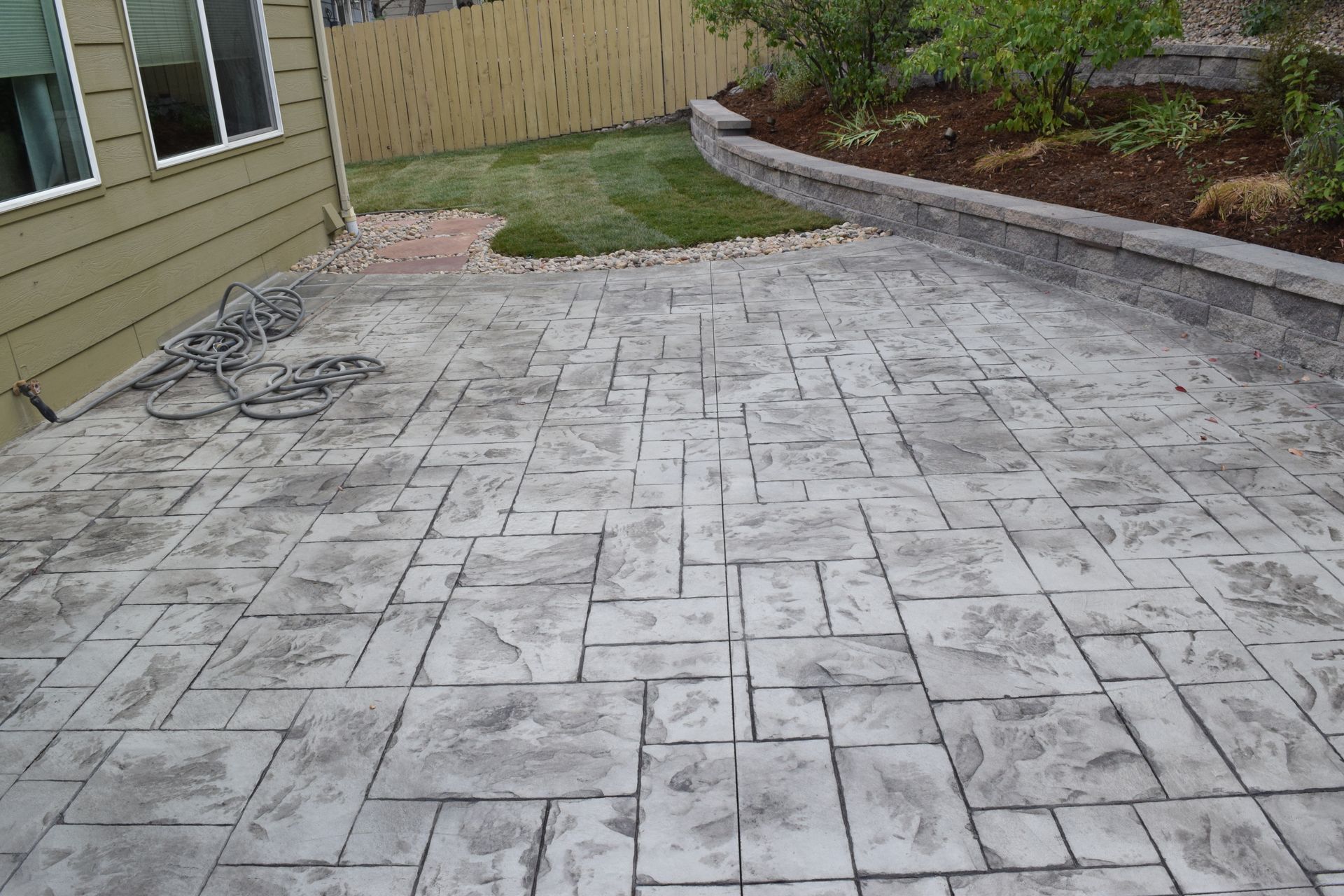 Concrete patio with a patterned design, next to a house and a retaining wall.