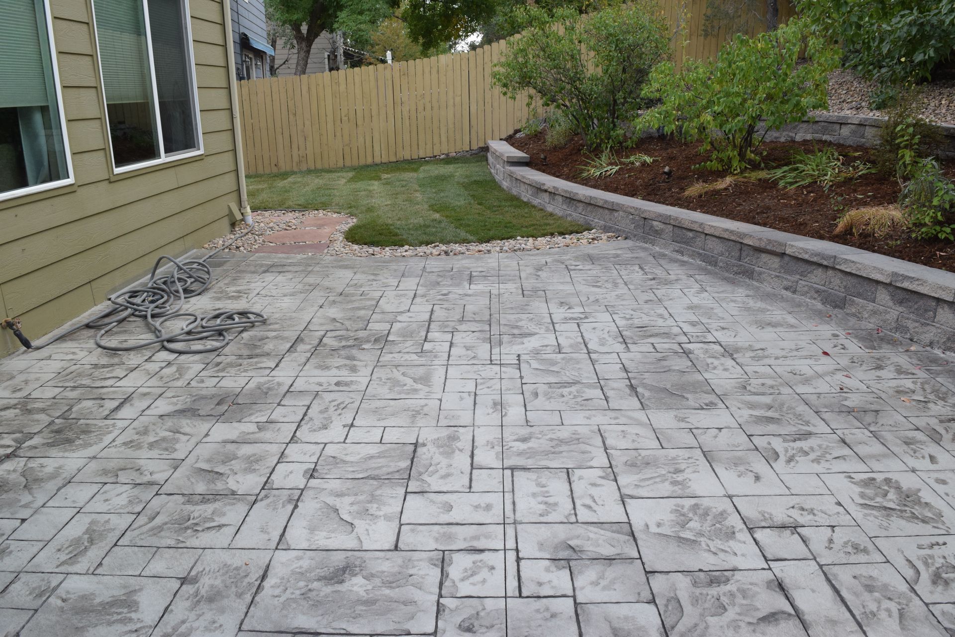 Stamped concrete patio, gray, next to a green lawn, wooden fence, and house.