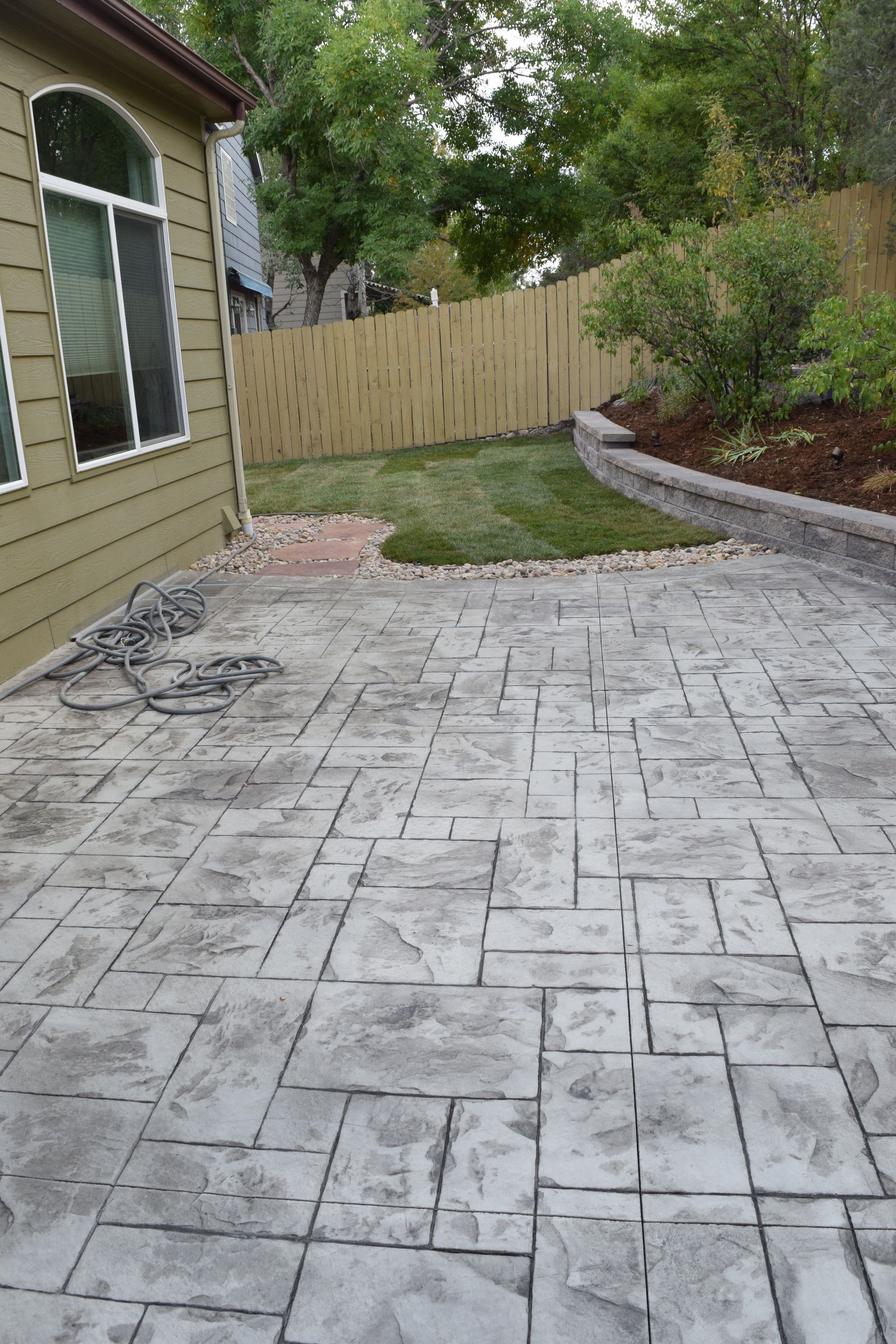 Stamped concrete patio next to a house with a small lawn, a fence, and landscaping.