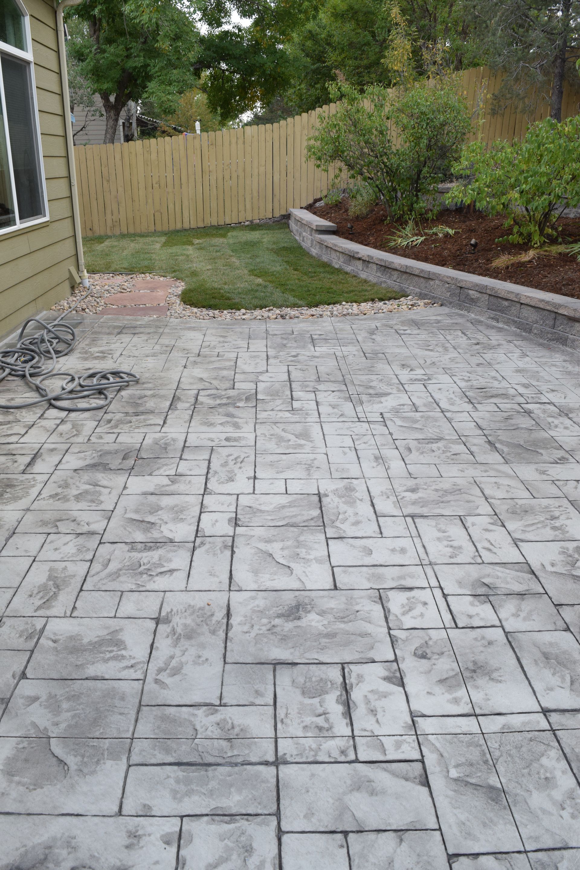 Grey stamped concrete patio with small lawn and flower bed. Fence in background.