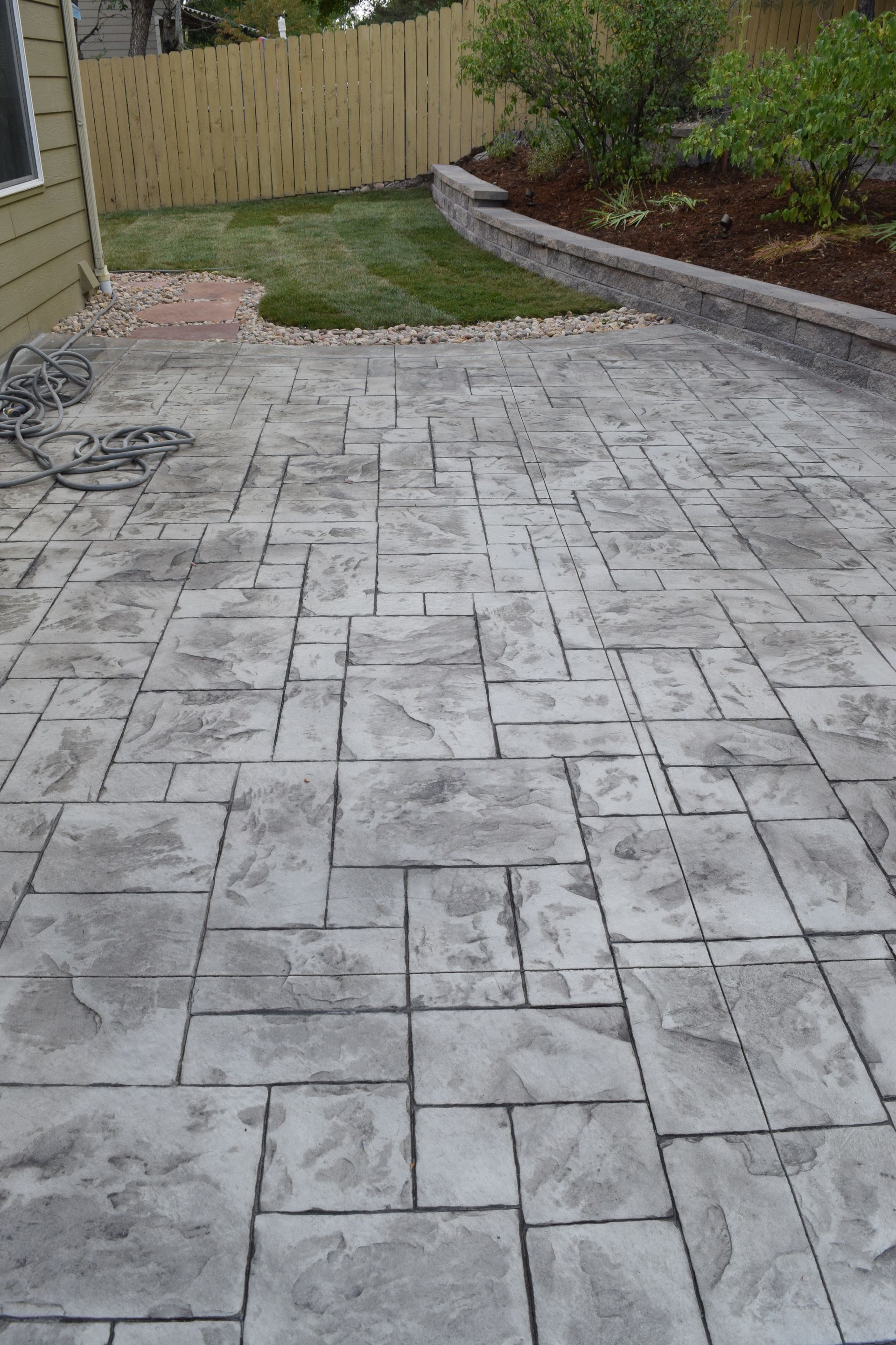 Gray stamped concrete patio with garden, lawn, and wooden fence.