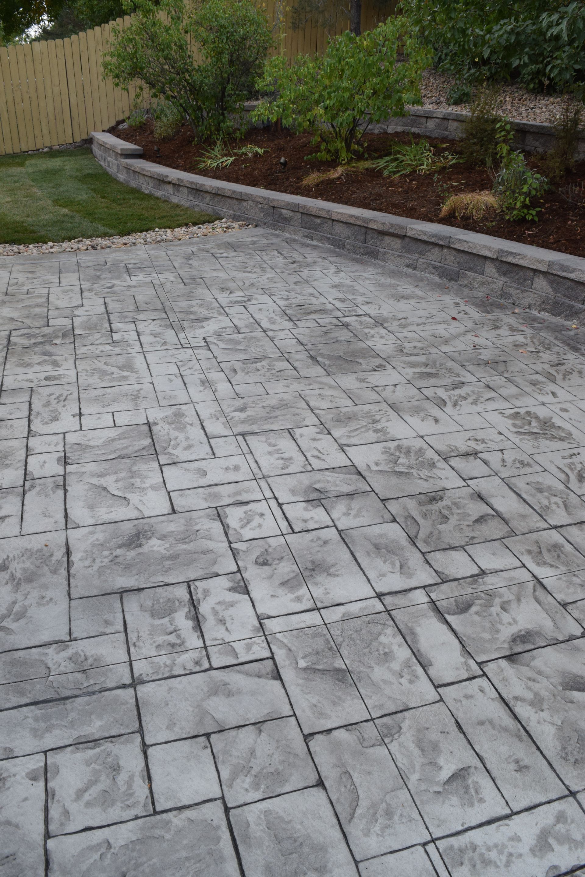 Gray stamped concrete patio leading to a tiered garden with greenery and a wooden fence.