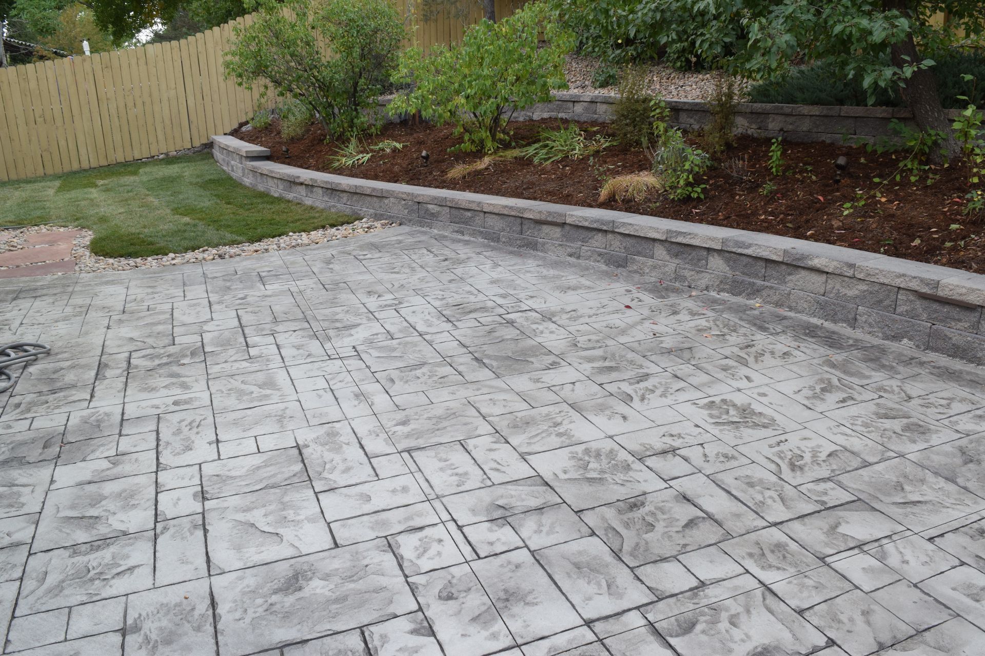 Stamped concrete patio with a retaining wall, grass, and a wooden fence.